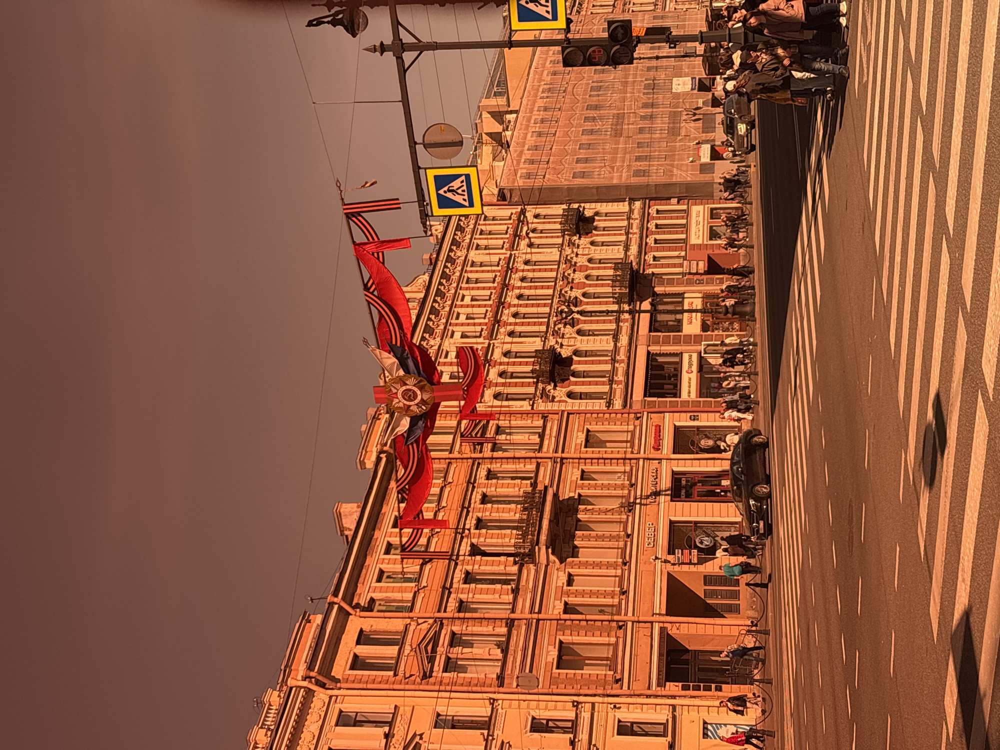 The image depicts a street scene in Saint Petersburg, Russia. A tall building with intricate architectural details is visible. The building is adorned with red banners, possibly for a celebration. Pedestrian crossing signs hang above. A crosswalk stretches across the road, where people are walking. A dark-colored car is parked on the street. The sky is overcast. The image is slightly tilted.