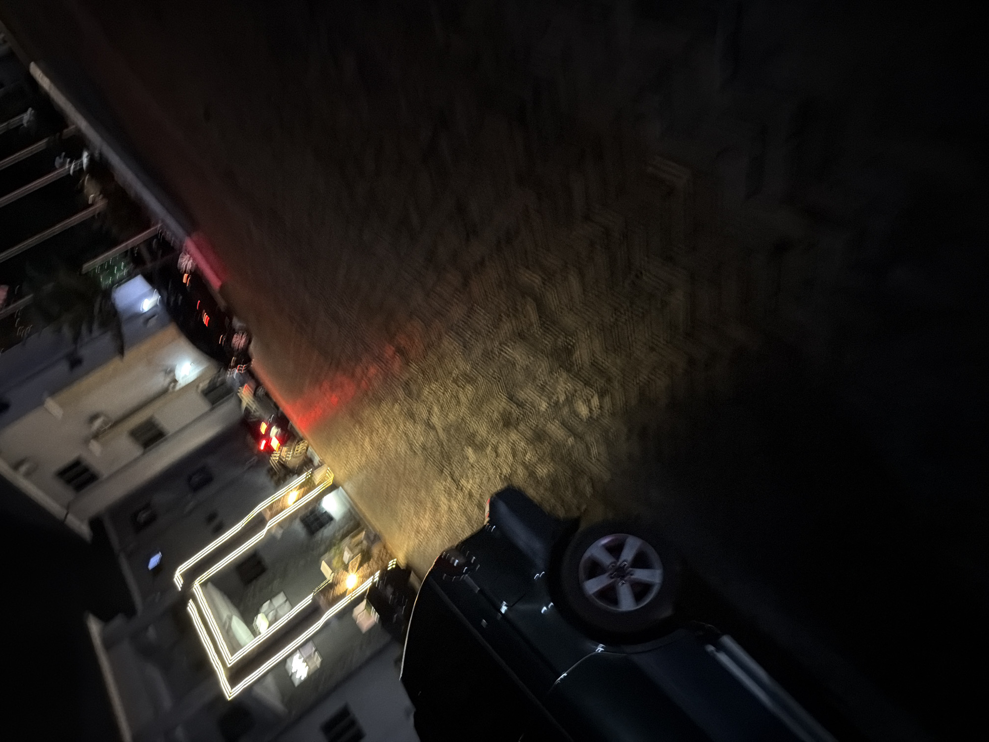 At night in Bukuru, Nigeria, a dark-colored car is parked on a cobblestone surface. The car's rear passenger-side wheel is visible. Behind the car, a building with illuminated windows and a geometric light installation is present. The scene is dimly lit, with reflections from the cobblestones and building lights casting glows.