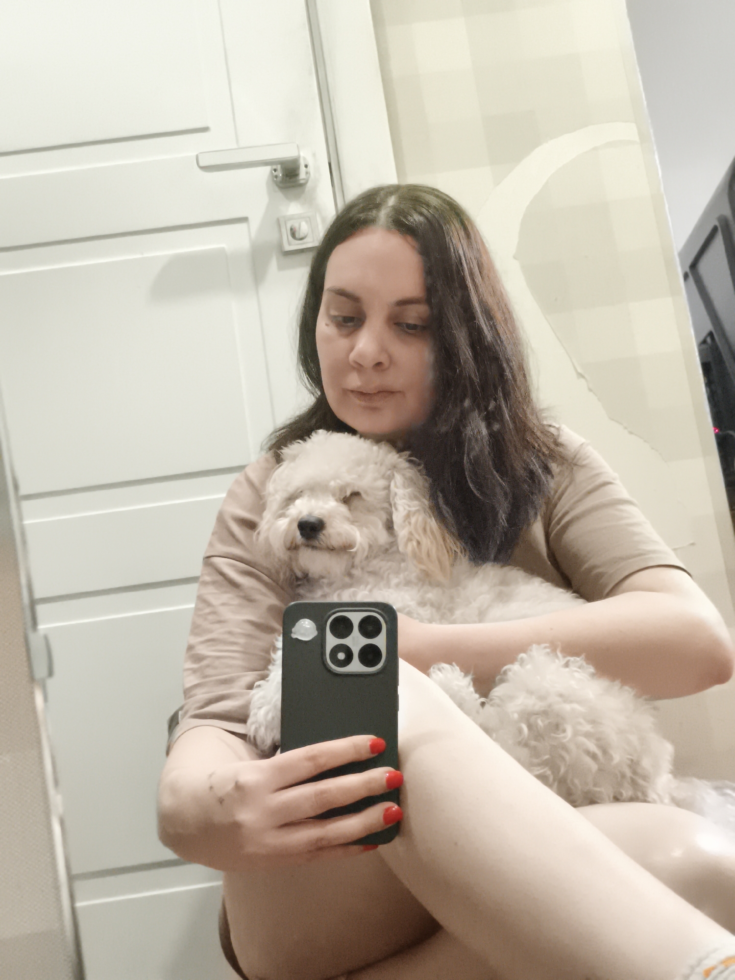 A woman with long, dark hair and fair skin is holding a white poodle while taking a selfie with a black iPhone. The woman is wearing a beige short-sleeved shirt and appears to be sitting. Her fingernails are painted red. The background includes a white door with a silver handle, and a wall with a light beige, patterned wallpaper. The image was taken in what seems to be Chaikovsky, Russia.