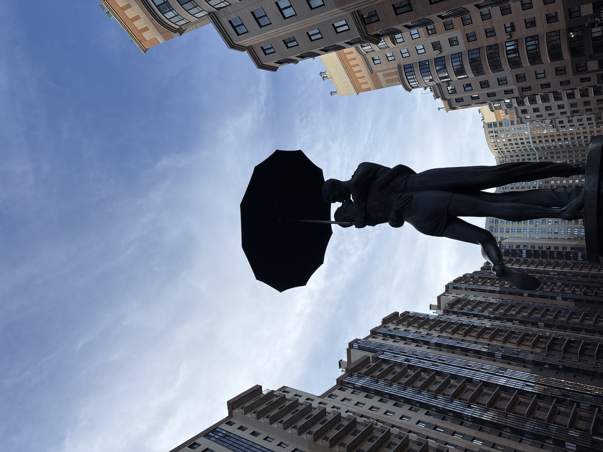 The image depicts a statue of a couple under an umbrella, framed against a backdrop of buildings and a blue sky with some clouds. The statue is dark, potentially bronze or a similar material, and is positioned so that the umbrella faces the top left of the frame. The buildings surrounding the statue have a modern architectural style with multiple floors and windows. The overall composition suggests the statue is located in an urban setting. The buildings' location and architectural style are not specific enough to confirm it is in Kudrovo, Russia.