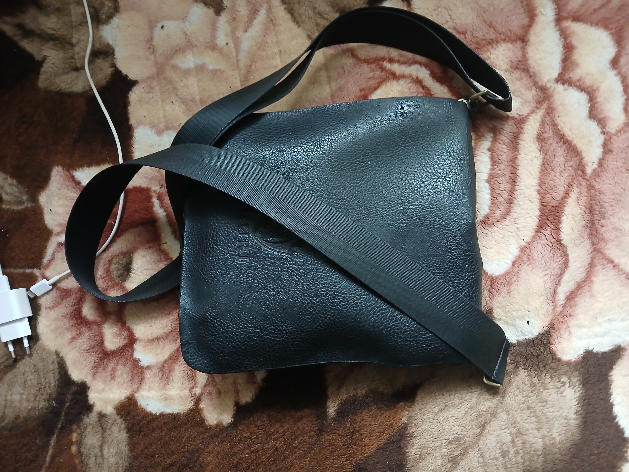 A black leather crossbody bag with a wide black strap is placed on a brown and pink floral patterned blanket. A white USB charger with a white cable is visible in the upper left corner of the image. The bag has a textured leather surface with a subtle embossed logo. The strap is diagonally positioned across the bag, with one end attached to the left side and the other extending out of frame to the right. The background blanket is soft and appears to be a fuzzy textile. The image is taken from a top-down perspective. There are no people or animals present. The location is specified as Hlushky, Ukraine, however, there are no visual cues in the image to confirm this location.