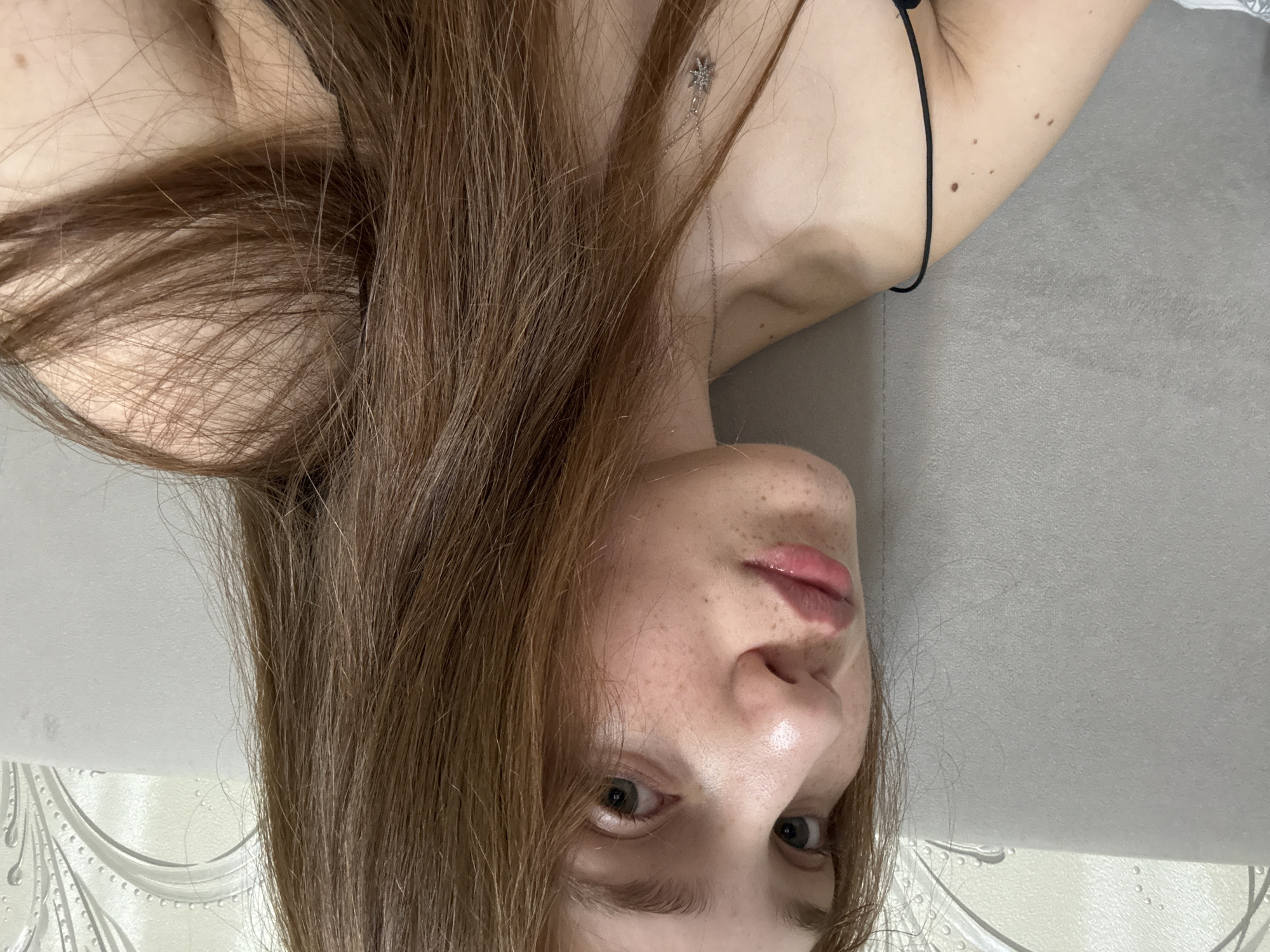 This is a close-up, self-portrait style photograph of a young woman with light skin, freckles, and long, reddish-brown hair. She is looking towards the camera with a neutral expression. Her lips are slightly parted and appear to have a gloss. She is wearing a thin black strap and a delicate silver necklace with a star-shaped pendant. The background consists of a textured grey surface, likely a headboard, and a light-colored wall with a decorative pattern. The lighting suggests an indoor setting. There is no indication of the location being in Naberezhnye Chelny, Russia.