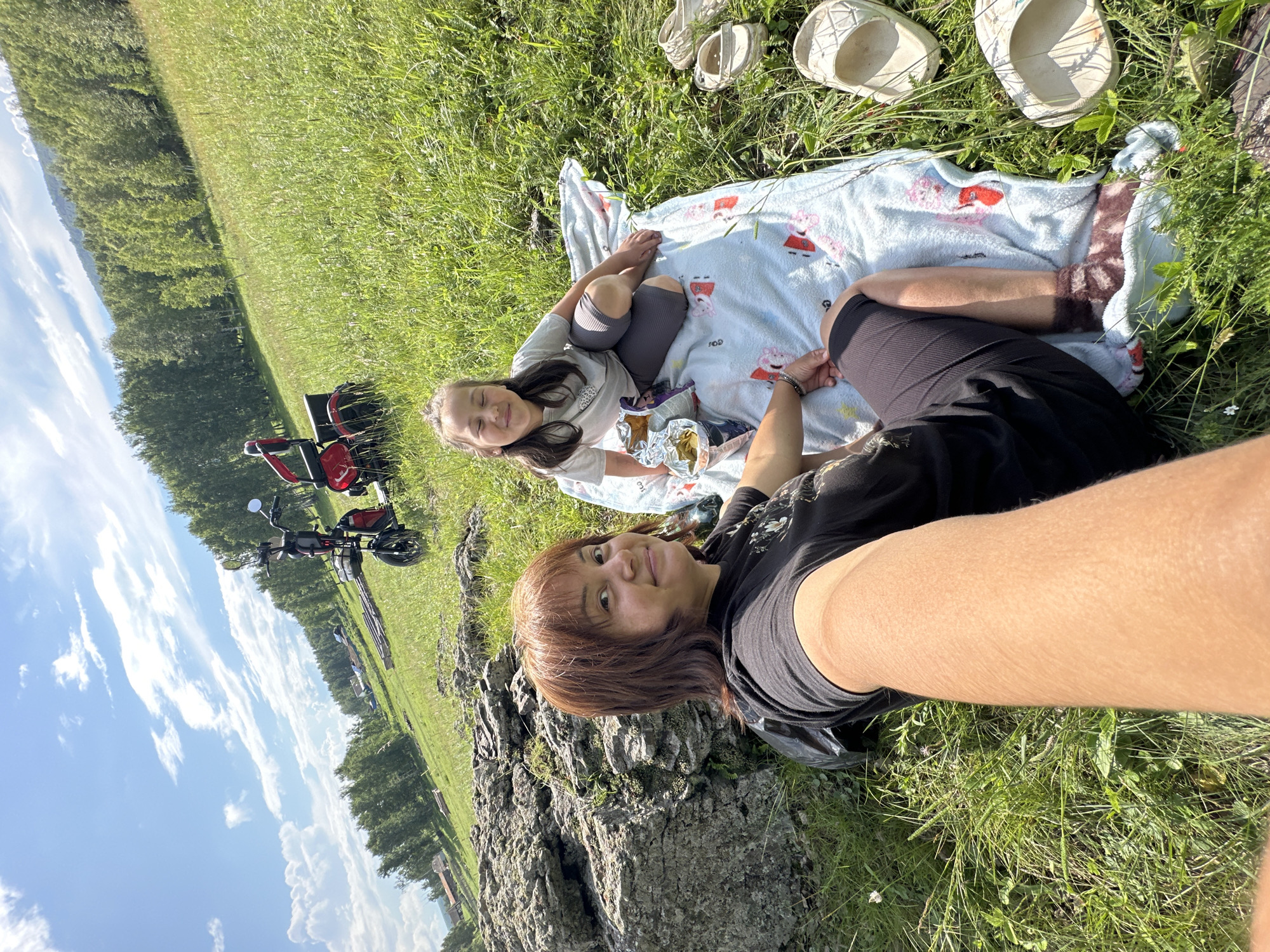 In this high-angle outdoor photograph, two individuals are enjoying a picnic on a grassy hillside. On the left, a woman with shoulder-length brown hair and bangs is seen from the chest up, taking a selfie. She is wearing a black t-shirt with a floral design. To her right, a young girl with her eyes closed and a slight smile is seated on a light blue blanket with a red and white pattern. She is wearing a light gray t-shirt and dark gray leggings. Beside her on the blanket is a bag of chips. In the background, a red and black electric scooter is parked on the grass. The environment appears to be rural, with a line of trees in the middle ground and a distant, treed mountain range under a bright blue sky with scattered white clouds. Several pairs of white clogs are scattered on the grass near the blanket. A large, textured rock formation is situated to the left of the woman. The overall scene depicts a relaxed summer outing.