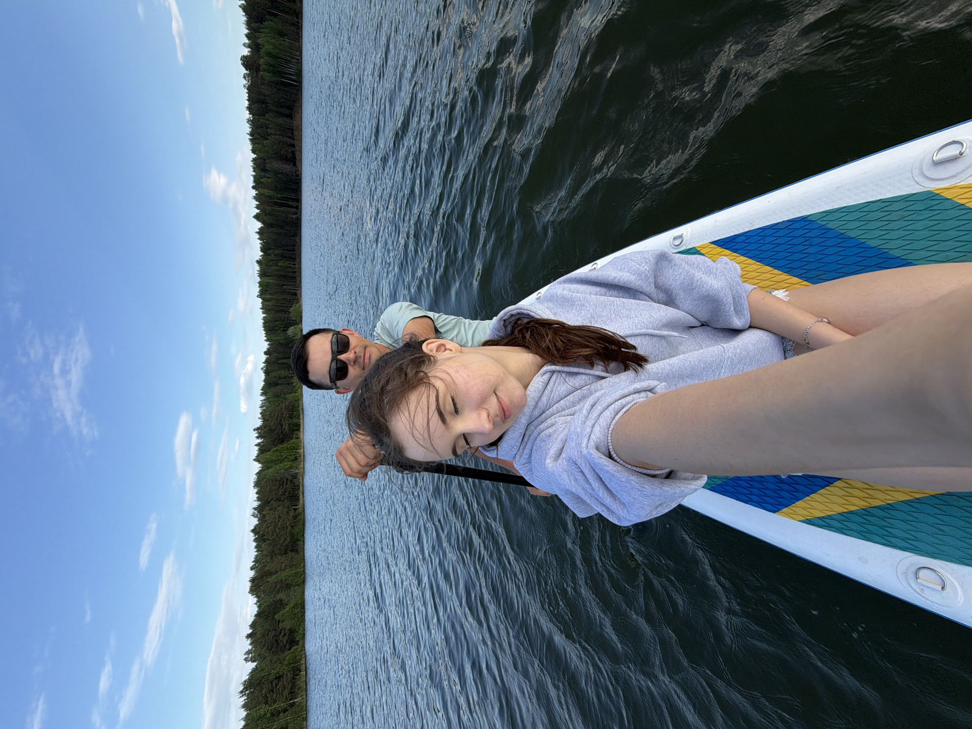 A man and a woman are lying on a paddleboard on a body of water. The man is wearing sunglasses and a light green shirt, and he is looking towards the camera. The woman is wearing a gray sweatshirt and has her eyes closed. The paddleboard is blue, yellow, and green. In the background, there is a line of trees and a blue sky with white clouds. The location is likely Selychka, Russia, given the request parameters. The couple seems to be relaxing and enjoying their time on the water.