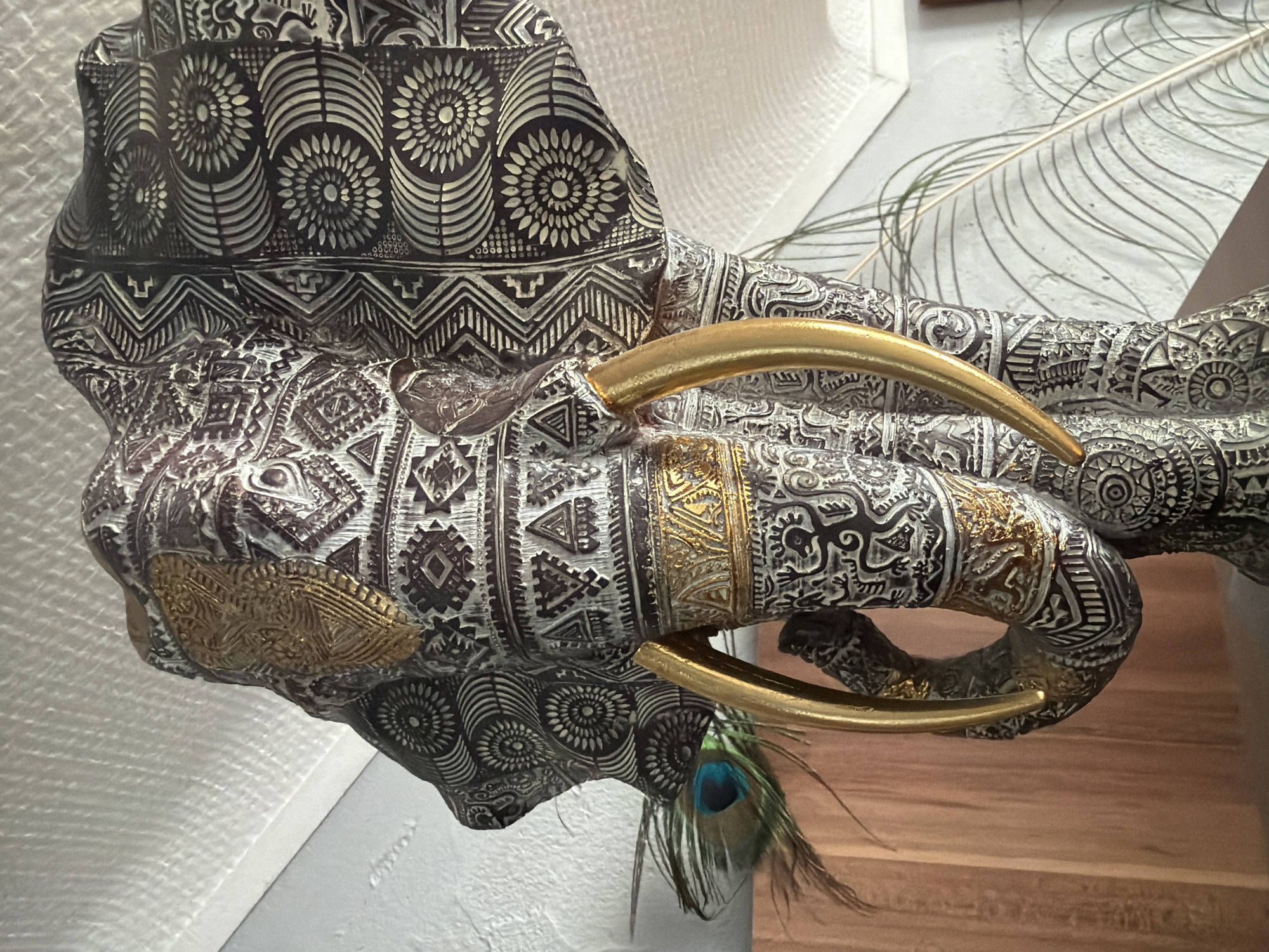 An ornate elephant head sculpture, intricately detailed with geometric patterns and gold accents, is positioned indoors. A peacock feather rests near the elephant's trunk. The background consists of a textured wall and a white ceiling. The sculpture appears to be a decorative item. No specific location in Esch-sur-Alzette, Luxembourg, can be determined from the image.