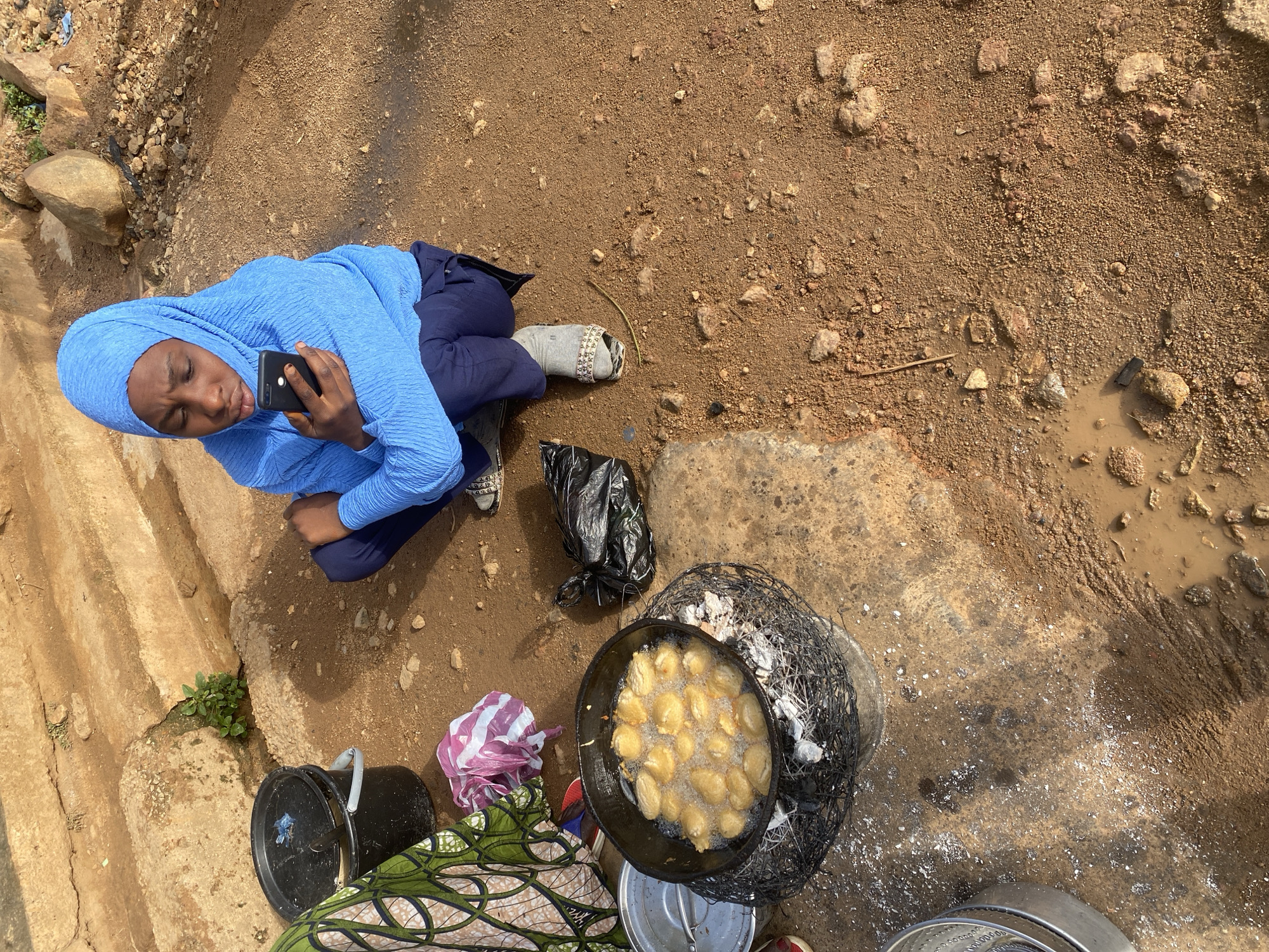 This overhead shot captures a young woman, likely in Jos, Nigeria, squatting on a dirt and concrete ground. She's wearing a bright blue hijab and matching tunic, along with dark pants and grey socks adorned with a pattern. In her hands, she holds a black smartphone, seemingly engrossed in its screen.
The scene appears to be an outdoor setting, possibly a market or roadside stall. To her right, a large black plastic bag lies on the ground. Further to the right, a black frying pan sits atop a makeshift wire grate over some smoldering embers, with numerous golden-brown fritters sizzling in hot oil. Next to the frying pan, a white and pink plastic bag is visible, and a patterned green cloth is spread out on the ground.
The ground itself is a mix of reddish-brown dirt, small stones, and patches of concrete. Puddles of murky water suggest recent rain or a water source nearby. A small green plant sprouts from a crack in the concrete on the left side of the image. The overall lighting suggests it is daytime, with clear weather conditions. The woman's posture and focus on her phone suggest a moment of personal time amidst her surroundings.
