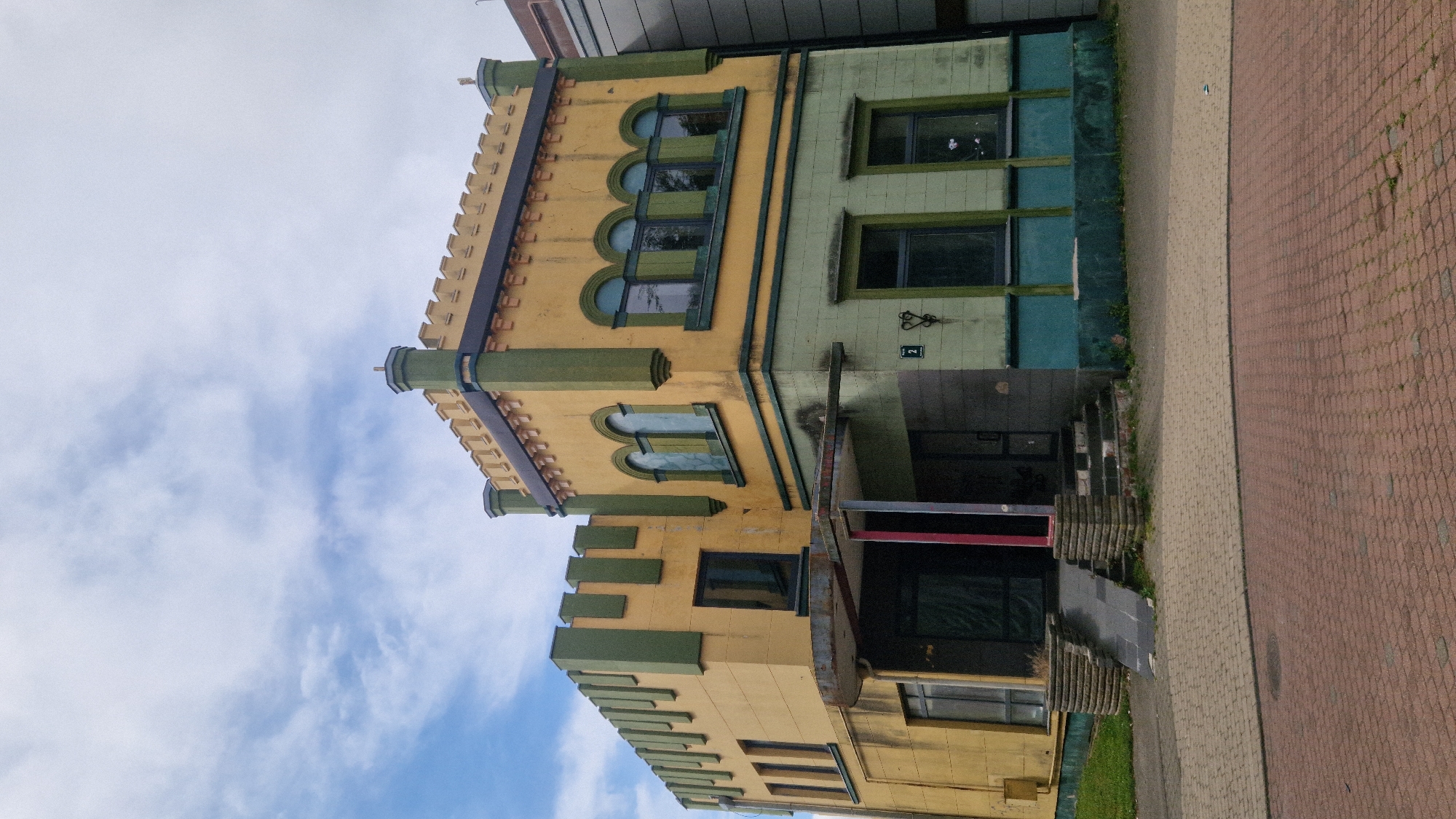The image depicts a two-story building with a distinct architectural style, possibly reminiscent of a castle or fortress, located in Liepaja, Latvia. The building's facade is painted in alternating bands of yellow and green, with crenellated decorative elements along the roofline. The lower level is predominantly green, with several rectangular windows. The upper level is yellow, featuring a series of arched windows. A covered entrance with a ramp and stairs leads to the main doorway. The building appears somewhat aged, with visible signs of wear and possibly neglect. The foreground shows a brick-paved walkway. The sky above is partly cloudy. There are no people visible in the image.