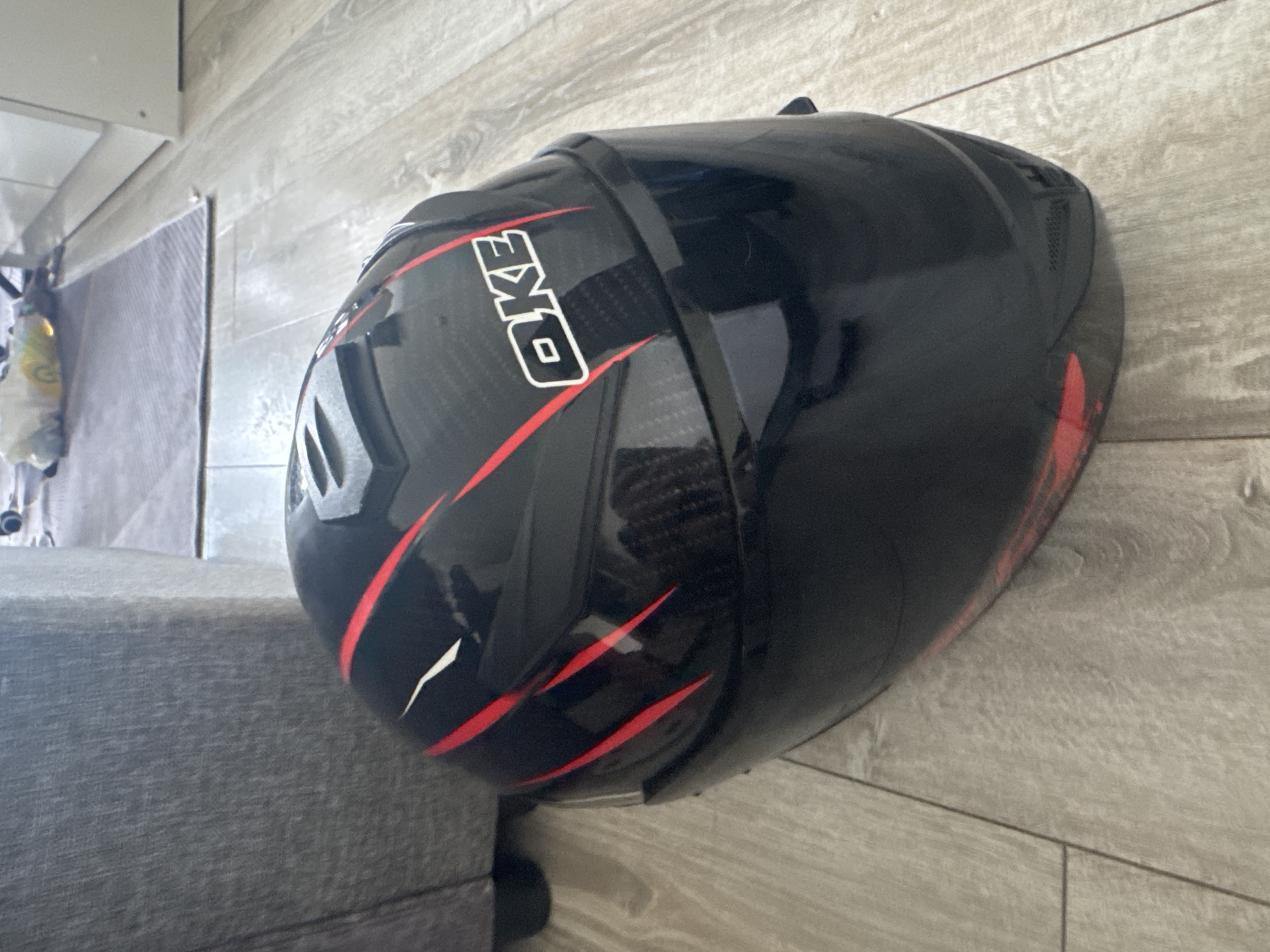 A black full-face motorcycle helmet with red racing stripes and a white "OKE" logo is lying on its side on a light-colored wooden floor. The helmet is positioned diagonally, with its front facing towards the right and slightly upwards. Behind the helmet, a portion of a gray upholstered piece of furniture is visible. In the background, out of focus, there is a glimpse of a white cabinet and a gray rug on the floor.