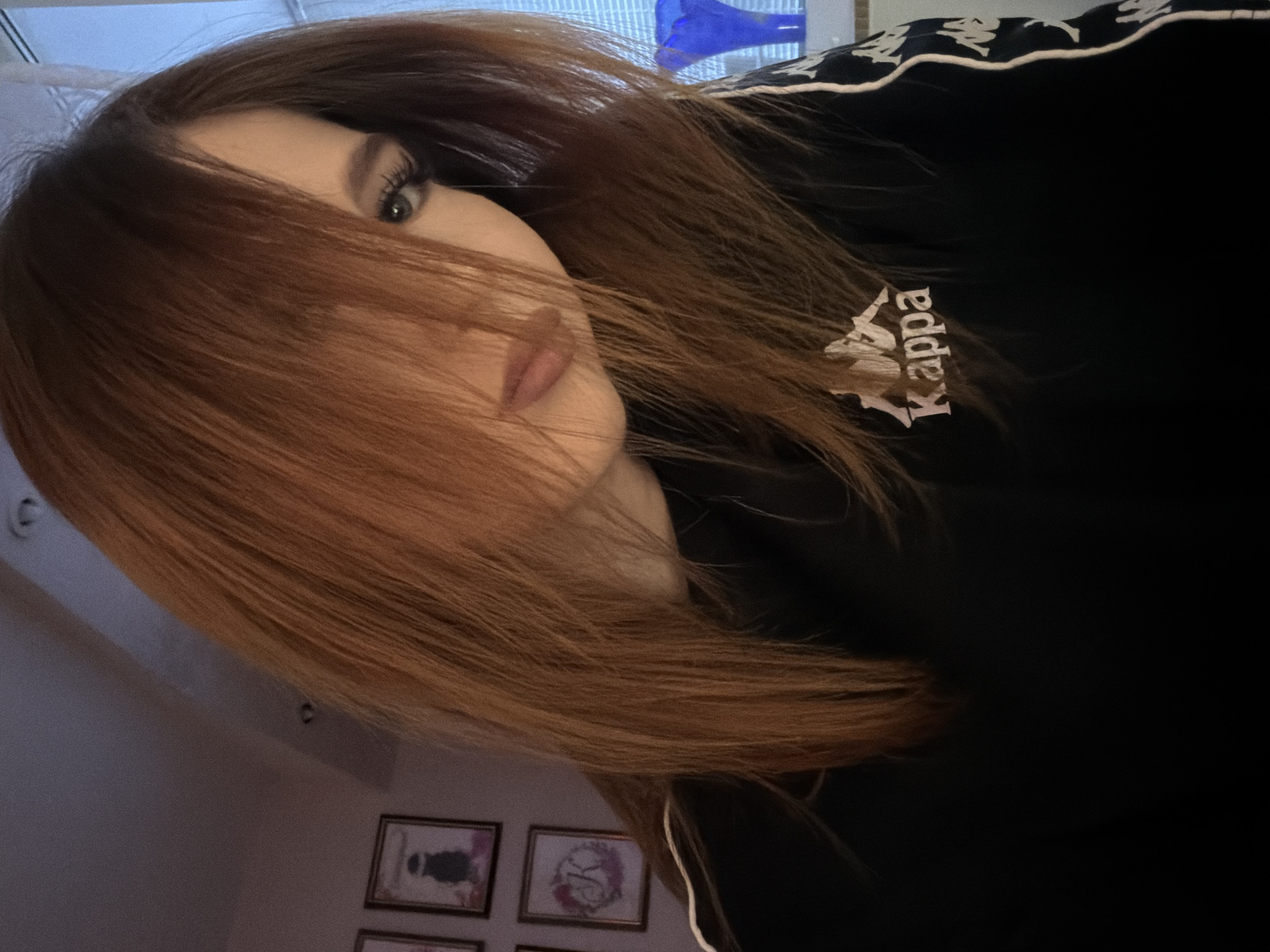 A photograph shows a person with long, auburn hair partially obscuring their face. The visible portion of the face includes a light-skinned complexion, one eye with dark eyelashes, and lips. The person is wearing a black Kappa-branded jacket with white detailing. In the background, partially visible, are framed pictures hanging on a light-colored wall. Based on the prompt's reference to "Noviy, Russia," the scene is assumed to be located there. However, without further visual context, the specific environment within the city cannot be determined.