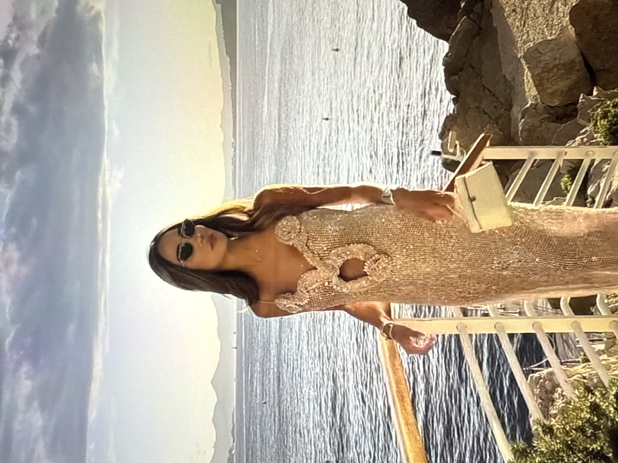 A woman stands on a balcony overlooking the sea, dressed in a shimmering gold dress and sunglasses. The dress features intricate detailing around the neckline and a cutout in the center. She holds a small white clutch bag in her left hand and rests her right arm on the balcony railing. The background consists of a body of water with distant mountains and a hazy sky. The scene appears to be a scenic coastal location. The image does not depict the city of Găvănoasa, Moldova.