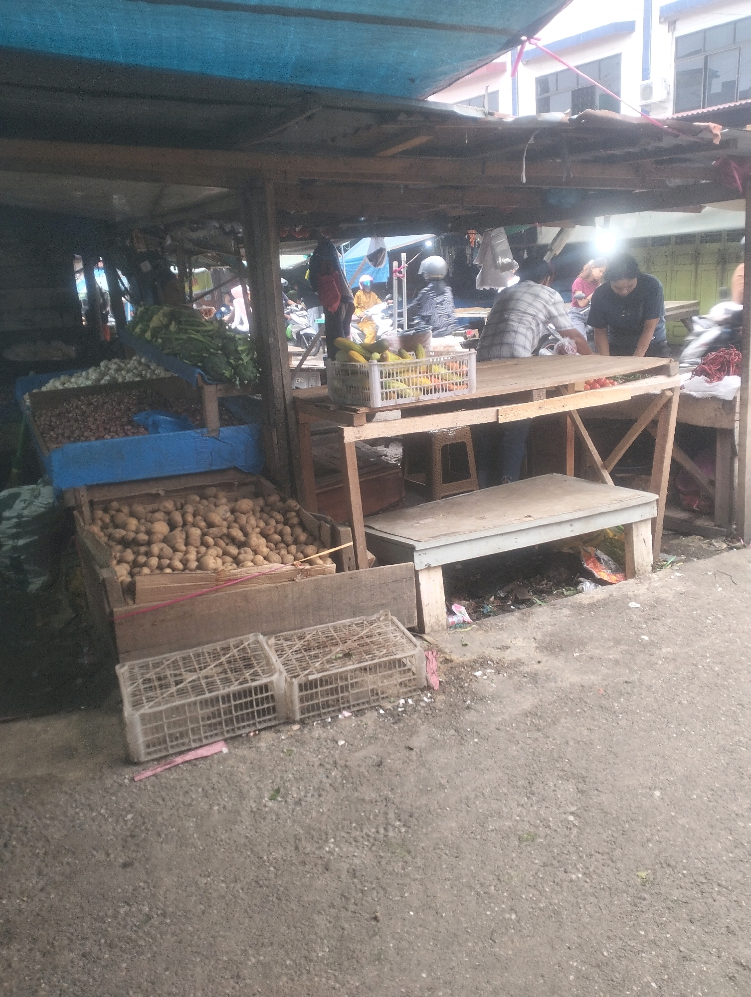 This image depicts an outdoor market stall in Balik Alam, Indonesia. A blue tarp covers the top of the stall, supported by wooden beams. Beneath the tarp, various produce is displayed. In the foreground, a wooden crate is filled with potatoes. To the left of the potatoes, another blue-covered crate holds onions. Further back, a white basket contains yellow fruits, possibly bananas. Behind the produce, several individuals are visible. One person, wearing a checkered shirt, is handling items on a wooden table. Another person, wearing a dark shirt, is also positioned behind the table. A third person, wearing a dark shirt and helmet, is seen in the background, operating a motorcycle. The market floor is covered with gravel and debris. The overall scene suggests a busy marketplace environment with vendors and customers interacting.