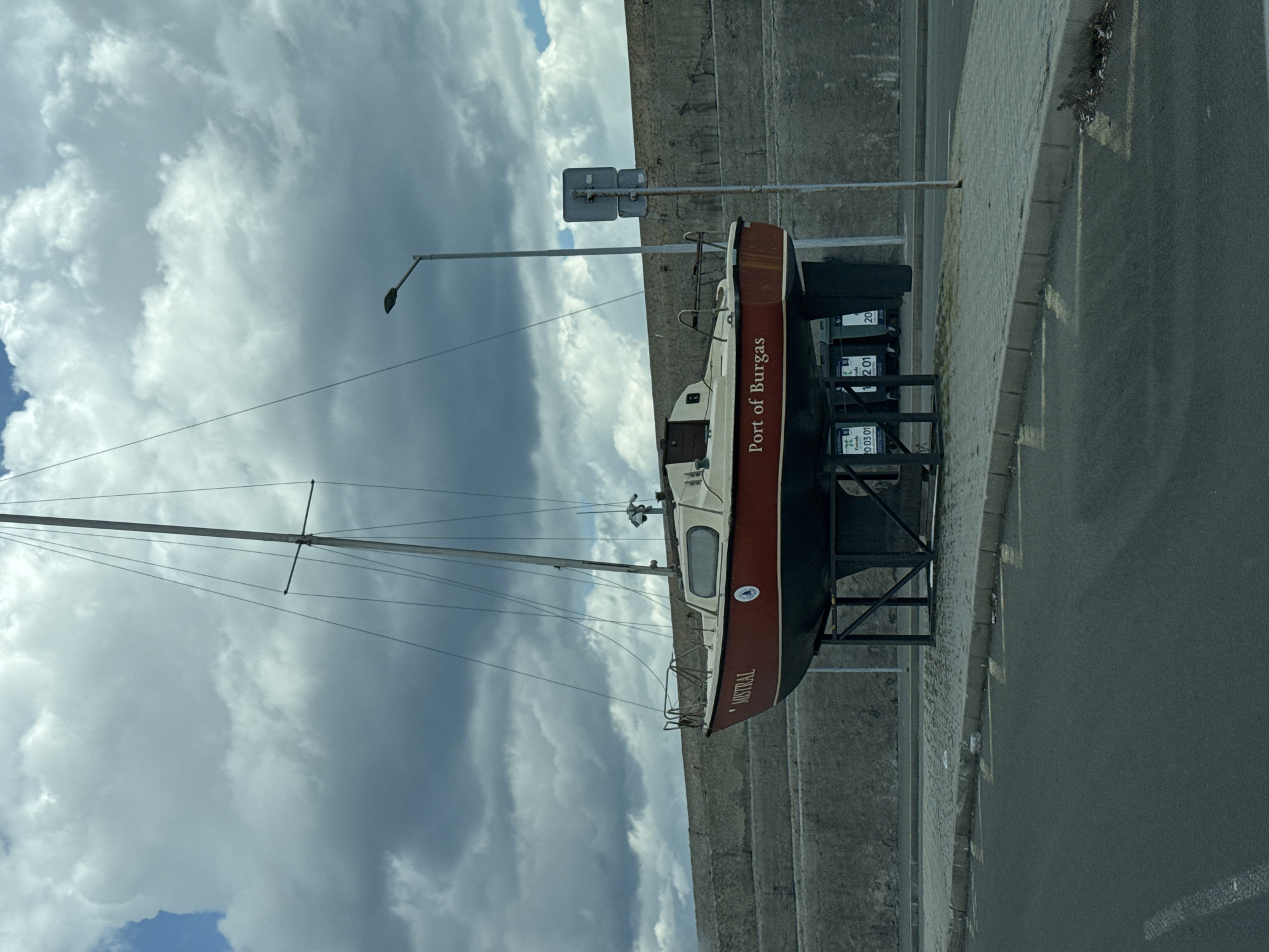 A sailboat, named "Mistral" and bearing the words "Port of Burgas," is mounted on a black metal frame on a gray brick sidewalk, near a street. In the background is a concrete wall and a cloudy sky. There is a light pole and a sign on the street.