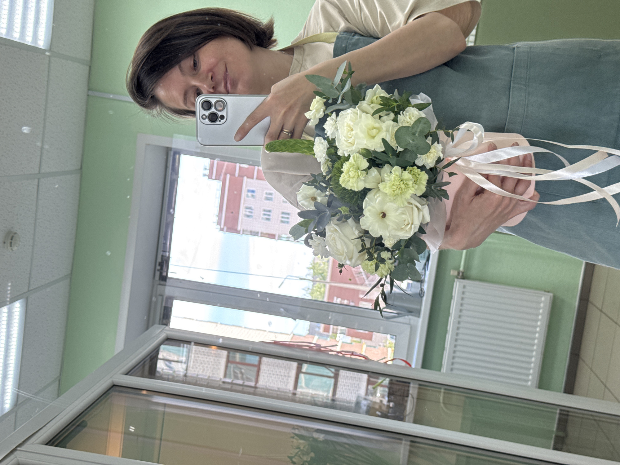 A woman stands in front of a large glass window or display case, holding a bouquet of white and green flowers. She is facing the window and taking a selfie with a silver smartphone. The woman has short, dark hair and wears a light-colored T-shirt under a blue-green apron. The bouquet is wrapped in a light pink material and tied with cream-colored ribbons. Outside the window, the reflection shows a cityscape with red brick buildings, likely indicating the location as Izhevsk, Russia. A white radiator is visible on the wall behind the woman. The ceiling has a grid pattern, suggesting the location is a commercial or office space. The window frame is white. The image is taken at an angle.