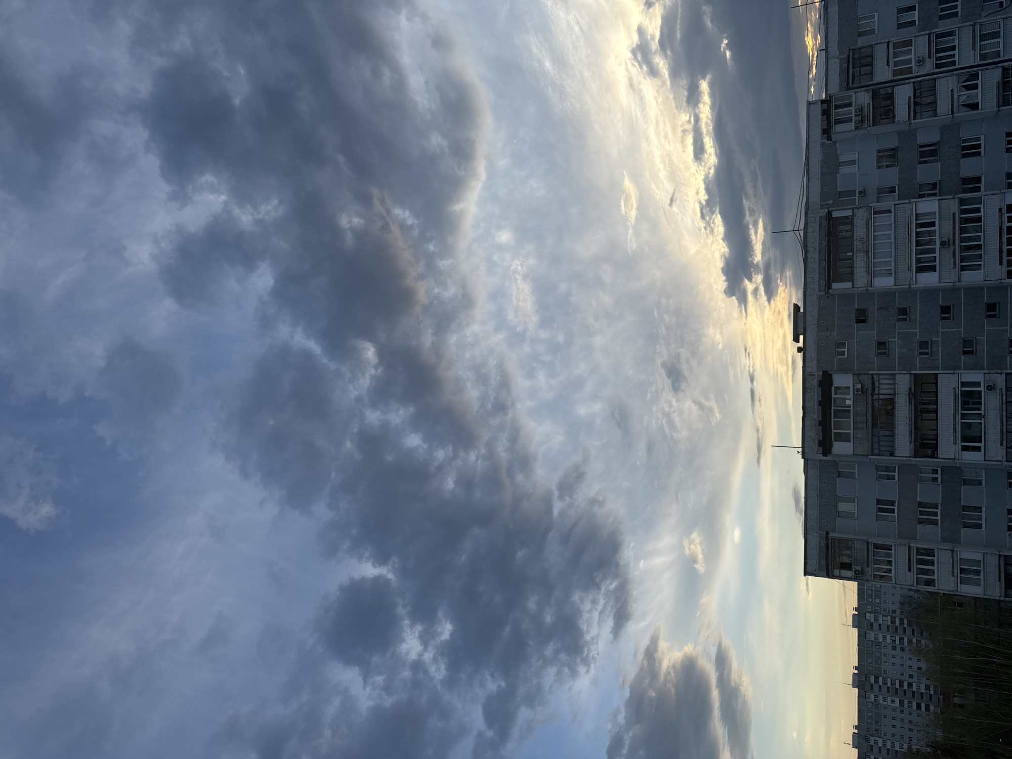 The image shows a cloudy sky above residential buildings in Kazan, Russia. The buildings are multi-story, constructed of gray concrete, and feature many windows and balconies. The sky is filled with a mix of dark and light clouds, with hints of sunlight filtering through. The image is rotated, presenting the buildings vertically on the right side. The environment appears to be urban, and the light suggests it might be evening or early morning.