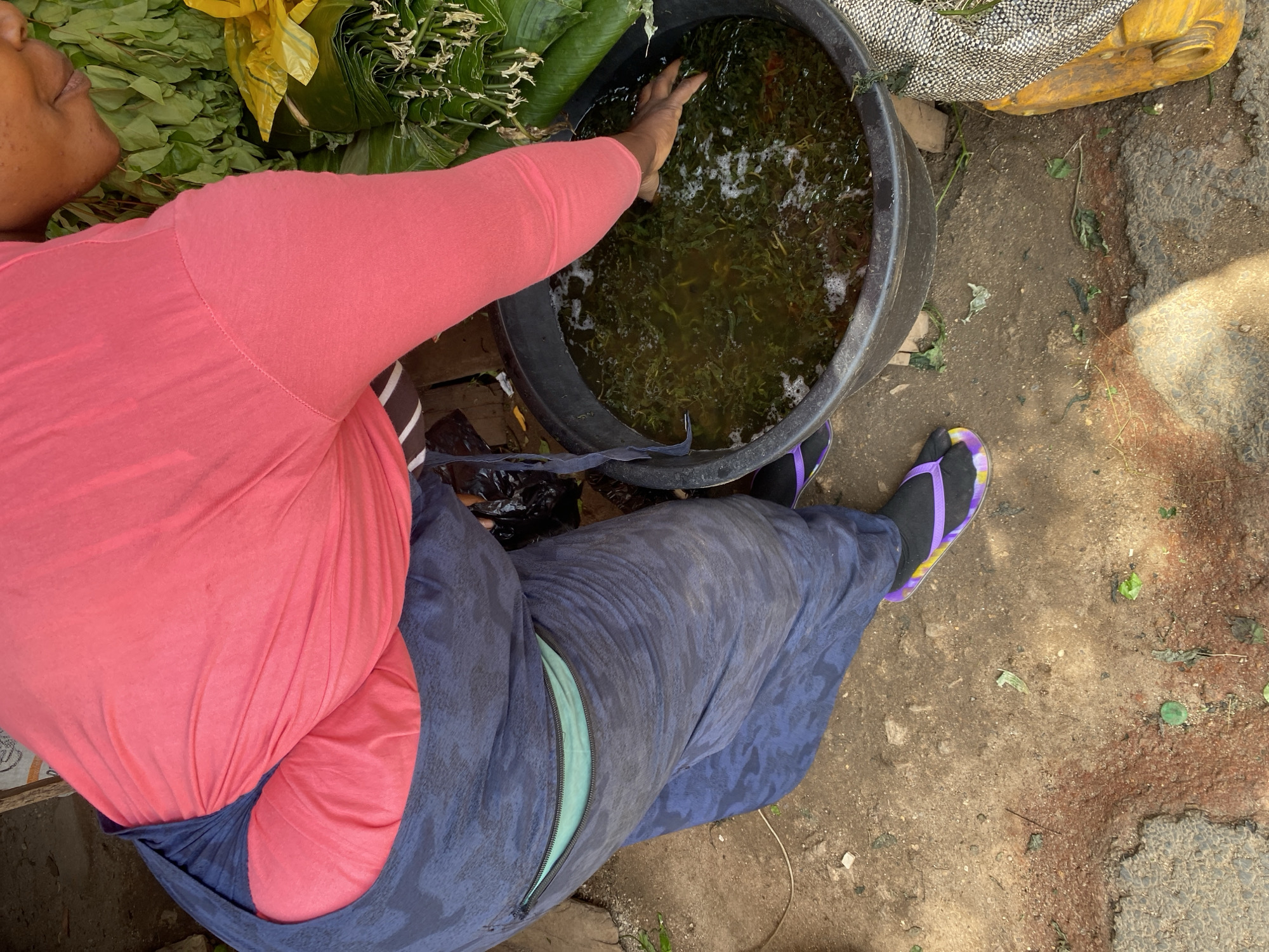 A woman, presumably a market vendor in Jos, Nigeria, is captured from a high angle. She is wearing a pink long-sleeved shirt and a dark blue patterned skirt with a bright mint green lining visible through a zippered opening. Her left hand is submerged in a large, black plastic basin filled with water and green leafy vegetables. The basin sits on a dirt and gravel surface, with some debris scattered around. To the right of the basin, a yellow object, possibly a container or sack, is partially visible, along with a bundle of green leaves wrapped in what appears to be a banana leaf or similar material. The woman is wearing black flip-flops with purple and yellow accents. The overall scene suggests a market setting, likely outdoors, given the natural ground cover and the presence of produce.