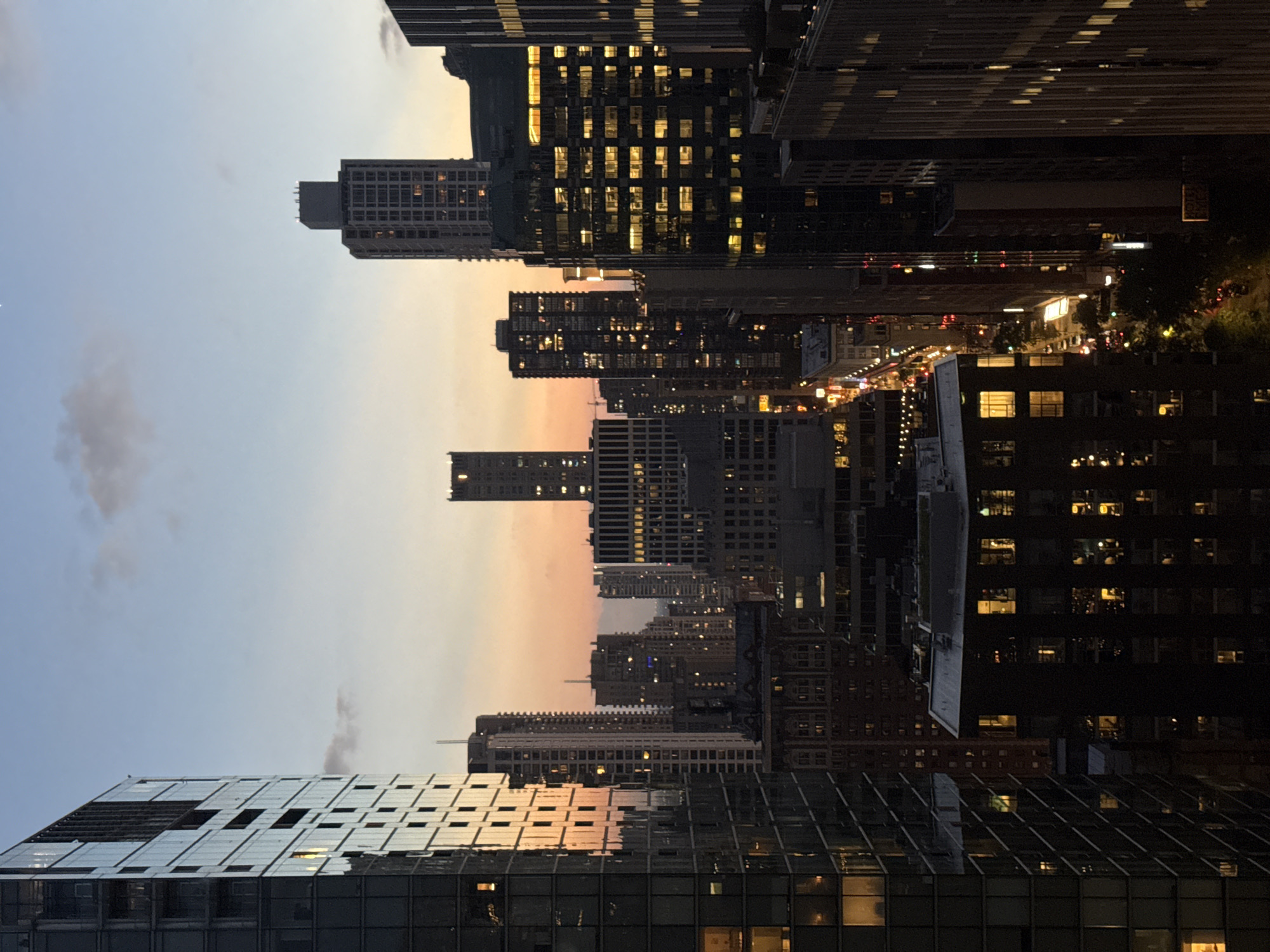 The image captures a dusk cityscape, likely Chicago, USA, with towering skyscrapers illuminated by interior lights. The sky displays a gradient of soft blues and oranges, indicative of sunset or twilight. A large, dark glass-fronted building dominates the left side of the frame, reflecting the warm hues of the sky and the lights of other buildings. To the right, a dense cluster of buildings of varying heights and architectural styles fills the midground and background. Many of these buildings have windows that glow with warm, yellow light, suggesting occupancy. The street level is partially visible in the distance, with hints of traffic and streetlights adding to the urban ambiance. The overall impression is one of a bustling metropolis settling into the evening.