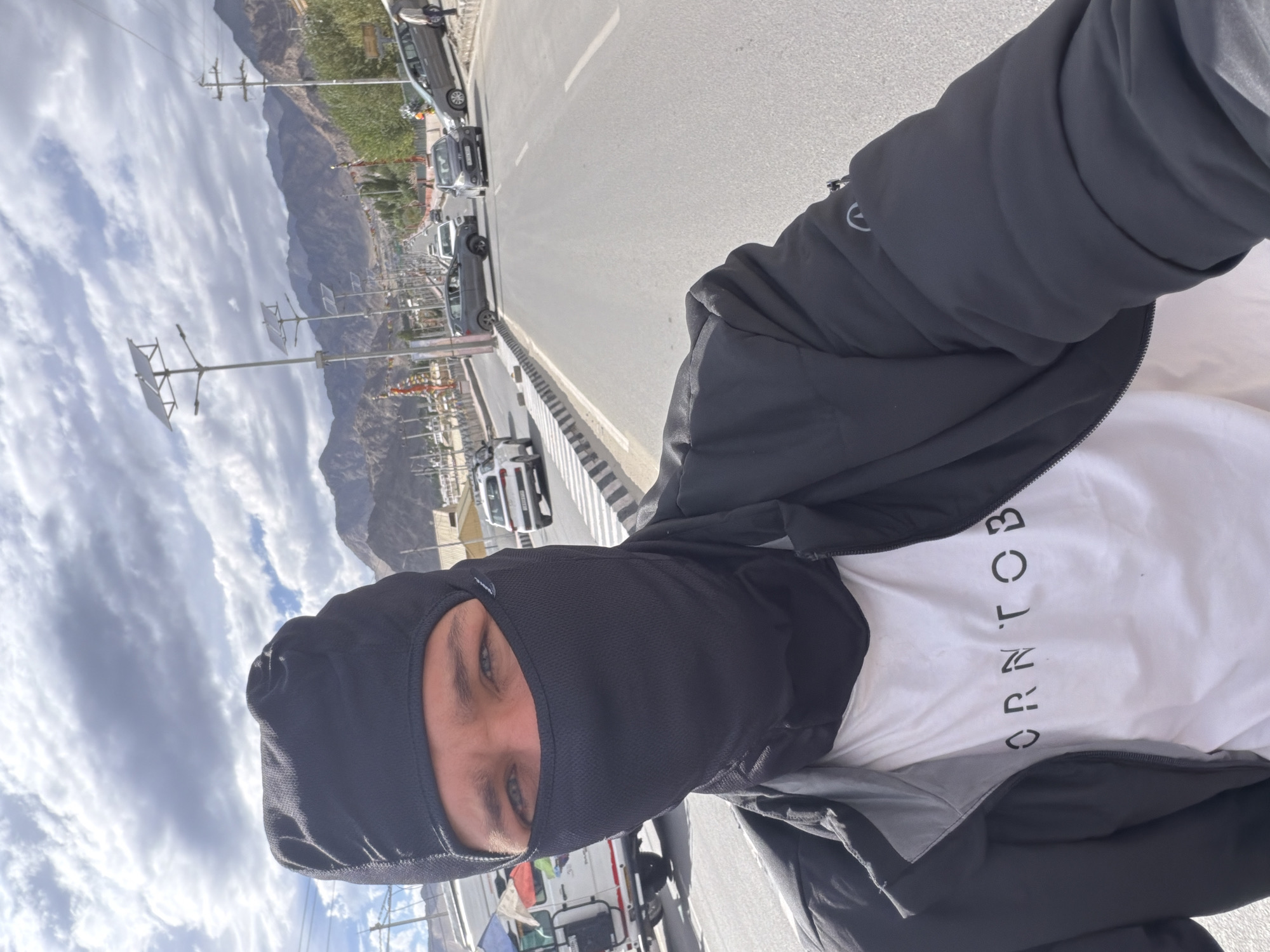 A close-up selfie shows a person wearing a black balaclava that covers their head and neck, with only their blue eyes visible. They are also wearing a white t-shirt with black text that reads "ORNT OB" and a black jacket. The person is on a road, likely in the city of Leh, India, as indicated by the background. The road has multiple lanes and is lined with streetlights featuring solar panels. Various cars, including a grey SUV and a white van, are present on the road. In the distance, mountains are visible under a cloudy sky. The scene suggests the person is traveling or in transit within a mountainous urban environment.