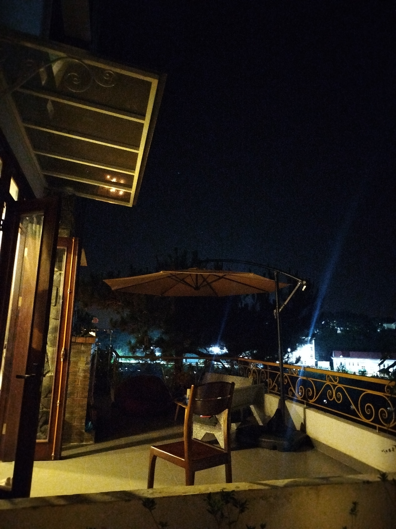 Night scene on a balcony in Lạc Dương, Vietnam. A wooden chair sits in the foreground near a stone ledge. Beyond the chair, a brown patio umbrella is partially visible, extending over a metal railing with decorative swirls. Behind the railing, city lights illuminate buildings and trees on a distant hillside. The balcony is connected to a building, with open wooden-framed glass doors visible on the left. An awning hangs above the doors, casting dim light on the balcony. The sky is dark.