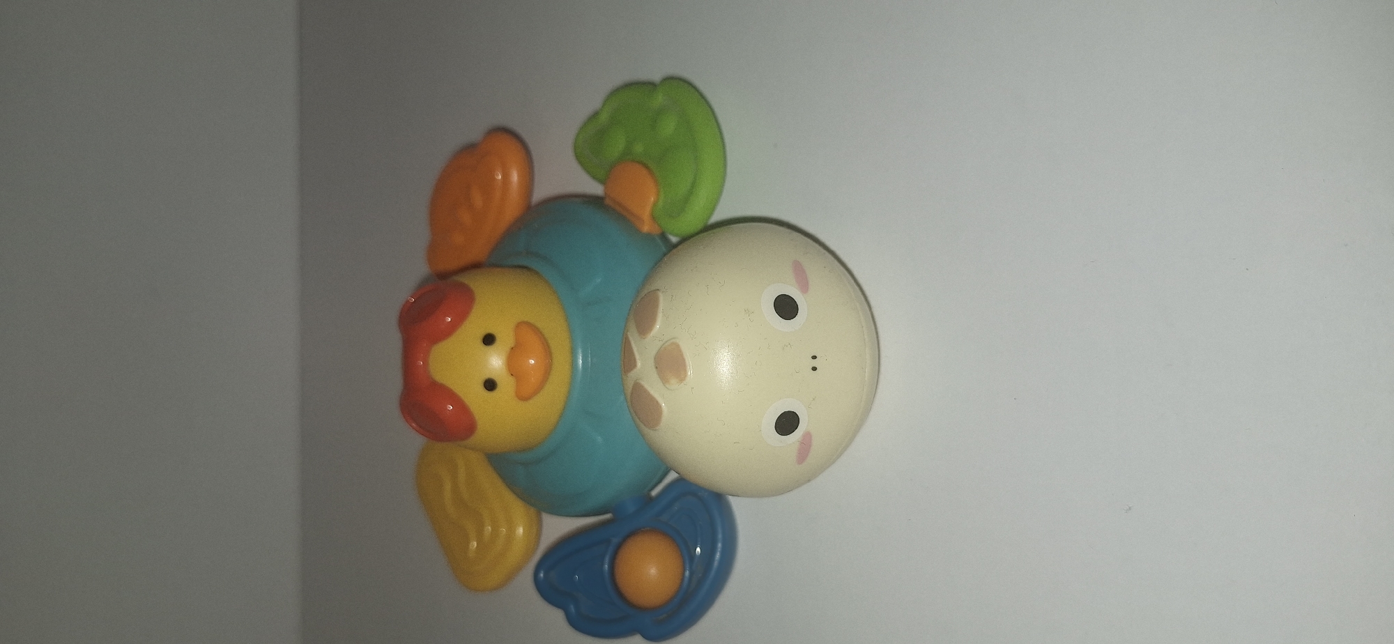 The image shows a colorful plastic toy with a yellow duck and a beige round body. The duck has a red comb, orange beak, and black dot eyes. It is attached to a turquoise body with orange and yellow wing-like attachments. In front of the duck is a beige round object with a face depicted by black dot eyes, pink circles for cheeks, and two small black dots for a nose. The object has a pattern of four light brown spots on its head. The toy is placed on a white surface against a light gray background. There is no indication of the location in Bila Tserkva, Ukraine.