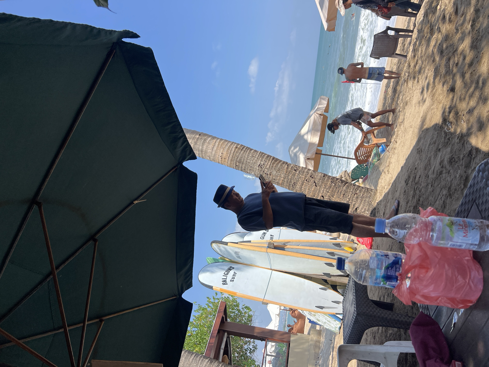 In Kuta, Indonesia, a man wearing a dark fedora and a navy blue t-shirt stands on a sandy beach, looking down at his smartphone. He is positioned to the right of a row of surfboards, which are leaning against a palm tree and a wooden structure. The surfboards are white with orange trim and have "BALIGUNS SURF" printed on them. To the left of the surfboards is a dark plastic table with two plastic chairs and two bottles of water. One bottle is a large clear bottle with a blue label, and the other is a smaller clear bottle with a red and white label. A red plastic bag is also on the table.
In the background, several beach umbrellas are set up, and people can be seen relaxing on beanbag chairs. The ocean is visible, with gentle waves breaking on the shore. Further out in the water, several surfers can be seen riding waves. The sky is a clear blue, with a few wispy clouds. The overall scene depicts a typical day at a popular tourist beach in Kuta.