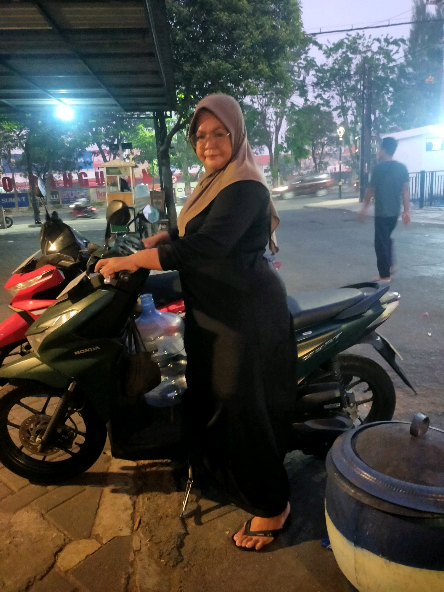 A woman wearing a hijab and glasses, dressed in a black abaya, stands beside two parked motorcycles. The woman is on the right, facing towards the viewer with her hands resting on the handlebars of a green motorcycle. A large blue water jug is strapped to the front of the green motorcycle. To the left of the green motorcycle is a red motorcycle. In the background, a man in a dark shirt and pants walks away from the camera, and a street with trees and buildings is visible. The scene appears to be outdoors, possibly a street or parking area, with some signage visible in the distance. The lighting suggests it is dusk. The presence of the word "HONDA" on the green motorcycle indicates it is likely a Honda brand vehicle. The context of "Surabaya, Indonesia" suggests this is a street scene within that city.