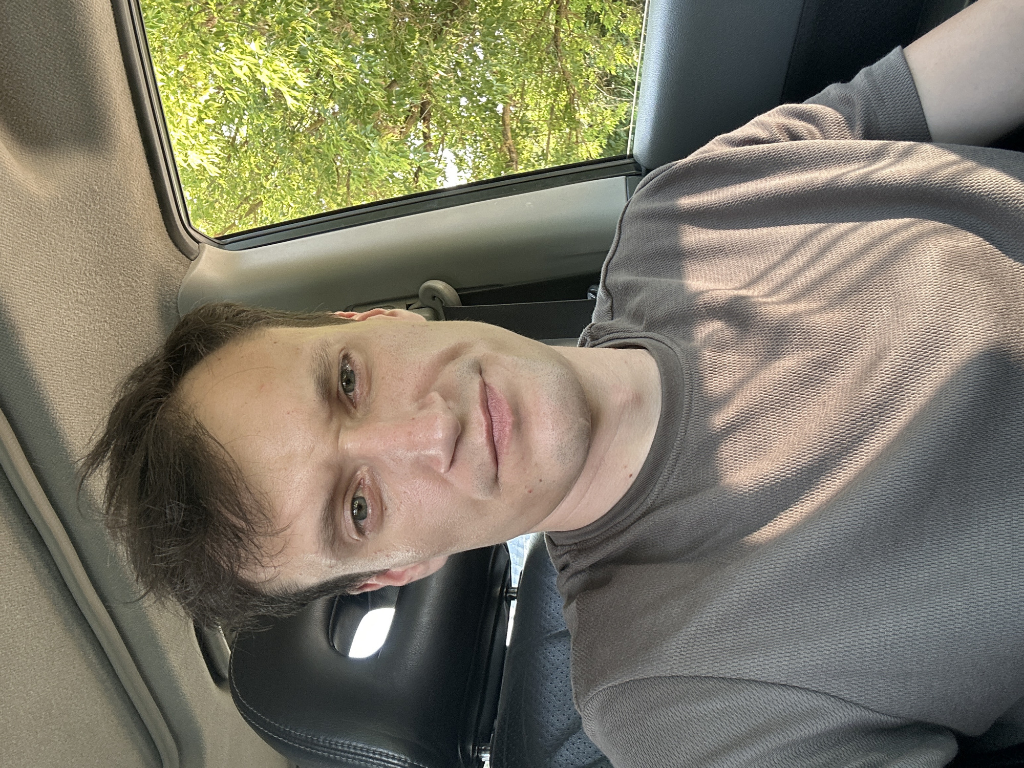 A man sits in the driver's seat of a car, his face turned towards the camera. He has short, dark hair and light-colored eyes. He is wearing a brown t-shirt. Sunlight filters through the car window, casting shadows on his face and shirt. Outside the window, lush green trees are visible, suggesting a natural or suburban setting. The interior of the car appears to be a typical car cabin with a visible headrest and part of the dashboard. The time of day appears to be late afternoon or early evening, indicated by the angle of the sunlight. There is no indication of the location within the city of Makiv, Ukraine.