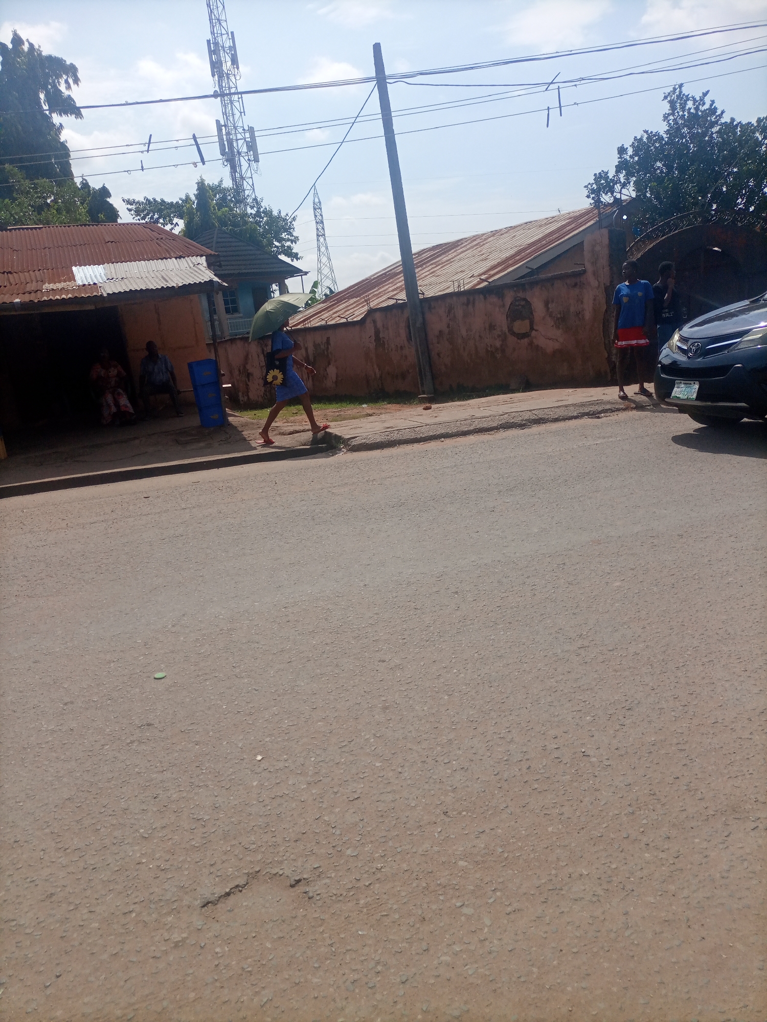 This is a street-level, eye-level, outdoor, daytime, medium-shot photograph taken in Nkpor, Nigeria. The foreground shows a cracked asphalt road with some small debris scattered across it. In the mid-ground, a woman is walking on a paved sidewalk, holding a green umbrella over her head. She is wearing a blue dress and red shoes. Behind her, a black Toyota SUV is parked on the right side of the frame. Further in the background, there are several buildings with corrugated iron roofs, a telecommunications tower, and trees. Two individuals are visible standing near the SUV. On the left side of the image, two people are seated outside a building. The sky is partly cloudy.