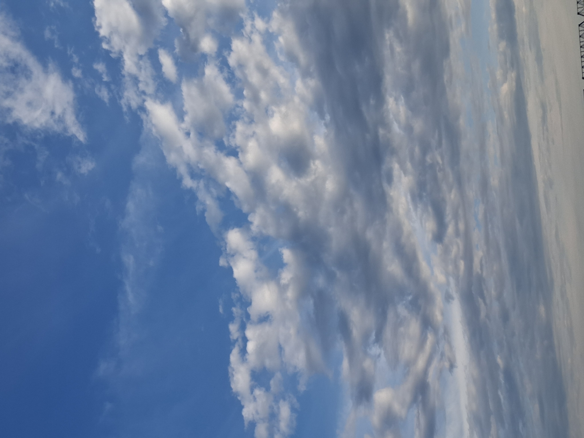 The image shows a cloudy sky with a blue hue. The clouds are varied in shape and density, with some appearing white and fluffy while others are gray and more solid. The lighting suggests it could be late afternoon or early morning. There are no visible people, subjects, or objects in the foreground or mid-ground of the image, only the sky and clouds. The bottom edge of the image shows a very faint hint of what appears to be a structure, possibly a bridge or some industrial construction, but it is too indistinct to identify. The image is a vertical, full-frame shot of the sky.