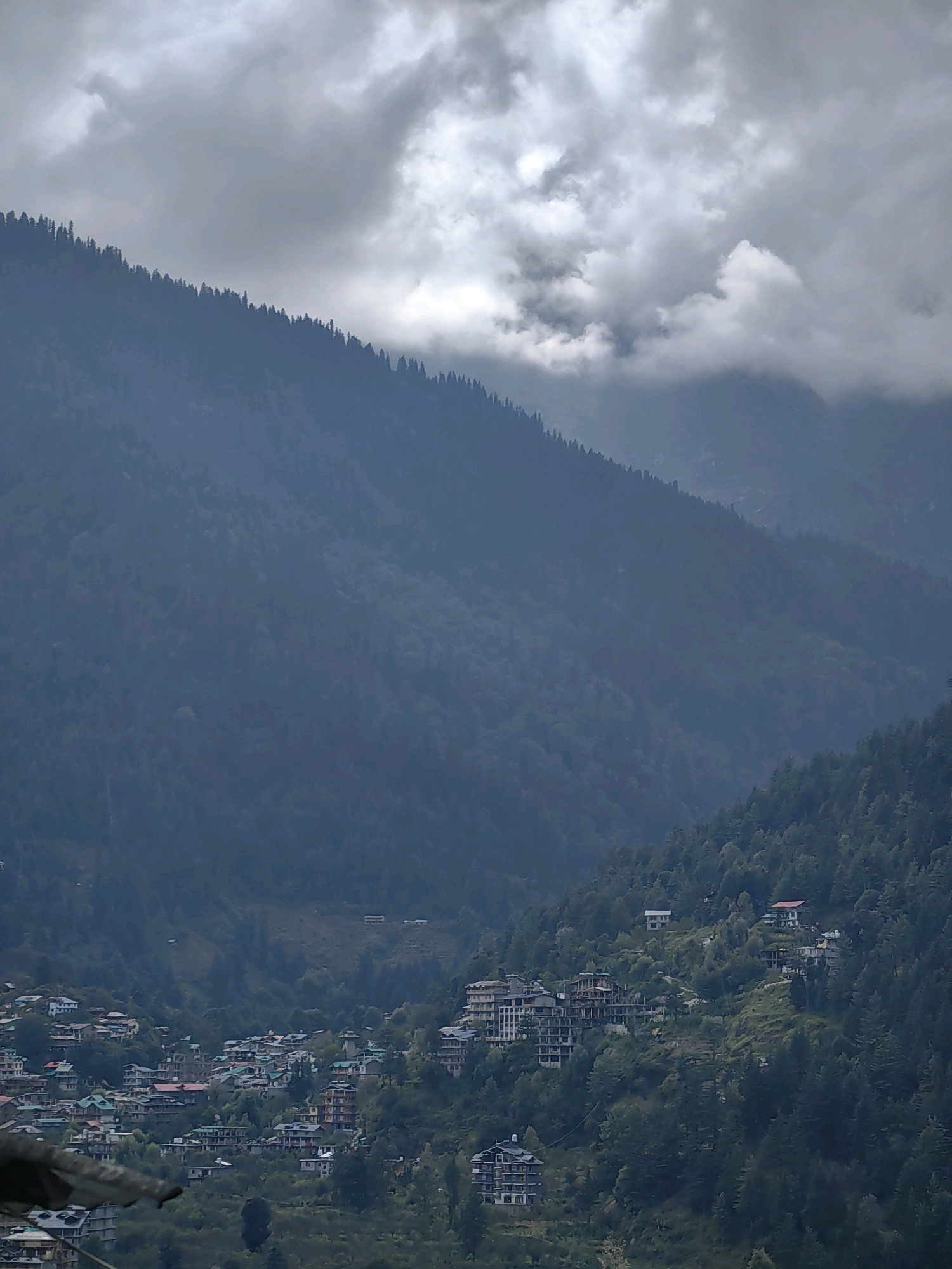 This is a high-angle, vertical, full shot of a town nestled within a steep, tree-covered mountain range under a cloudy sky. The town, identified as Vashisht, India, features a dense cluster of multi-story buildings, many with colorful roofs, scattered across the hillside. The buildings are a mix of architectural styles, with some appearing to be hotels or guesthouses, evident from their size and balconies. Dense evergreen trees cover the mountainsides, creating a lush, green backdrop. The sky is heavily overcast with dramatic, dark gray clouds, suggesting an impending storm or recent rain. The overall impression is one of a remote mountain community in a dramatic, natural setting.
