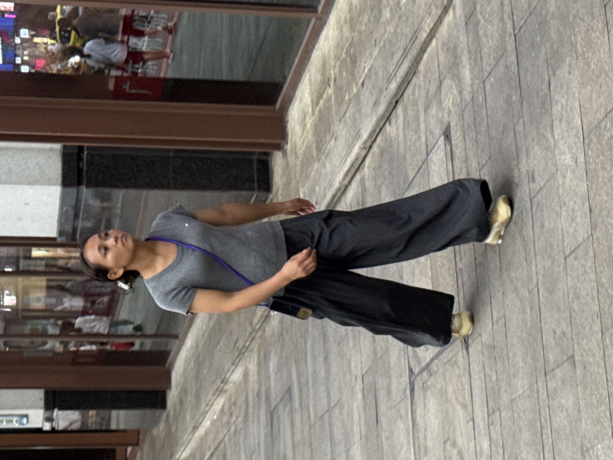 A woman walks on a paved sidewalk in Yuzhong District, China. She is wearing a grey t-shirt, black wide-leg pants, and yellow sneakers. A dark purple strap from a bag is visible across her body. The sidewalk is made of grey rectangular stones. In the background, large windows of a building reflect the street scene. The reflection shows people walking and a yellow car.