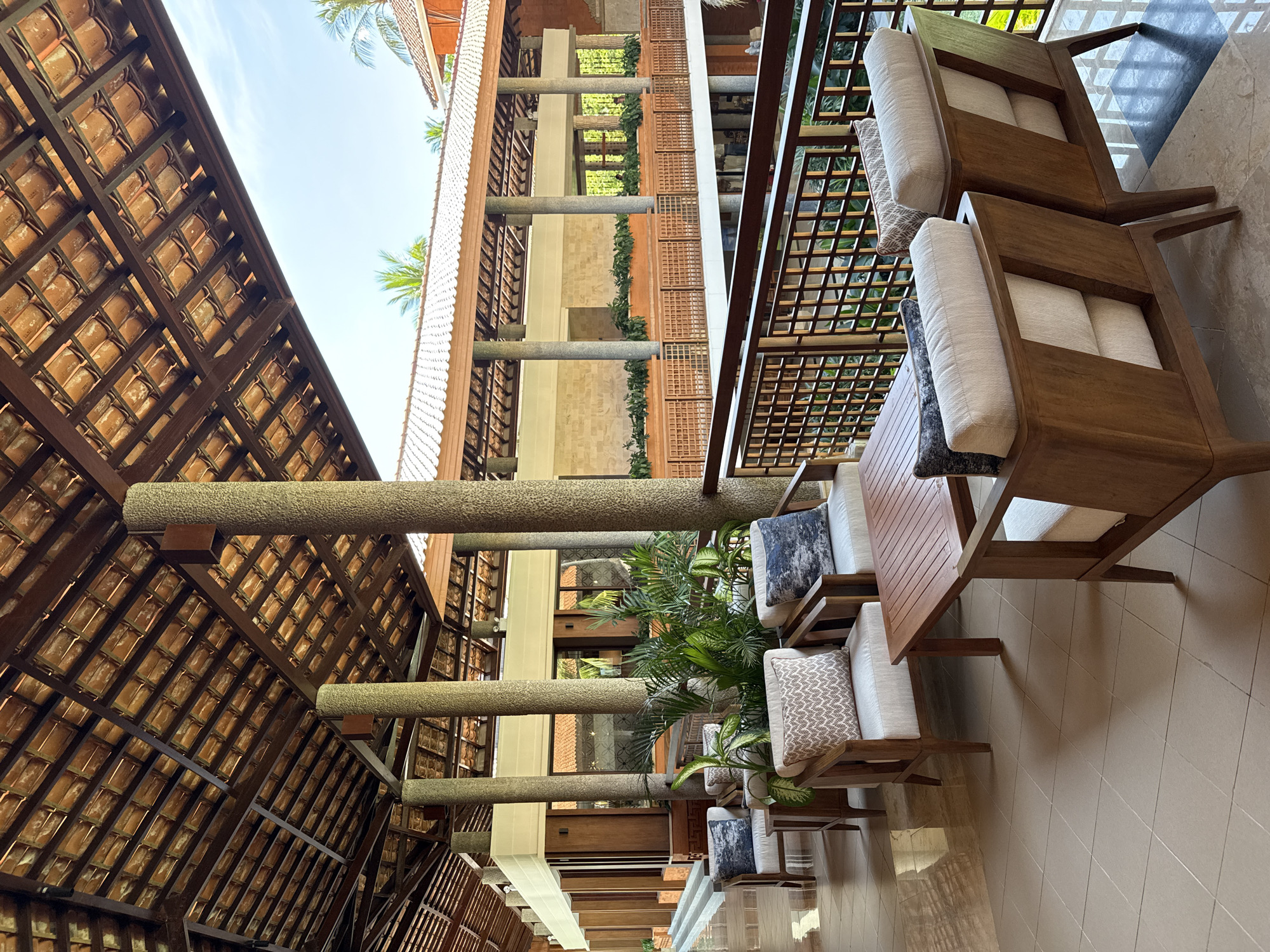 The image depicts a covered outdoor patio area in Kuta Selatan, Indonesia. The space is furnished with a series of wooden chairs with light-colored cushions and patterned throw pillows. A small wooden table sits between two of the chairs. The patio is part of a multi-story building, and a brown railing with a latticed design separates the patio from the open area below. The building features support columns made of textured stone, and the roof is covered in terracotta tiles. The overall setting evokes a tropical resort atmosphere.