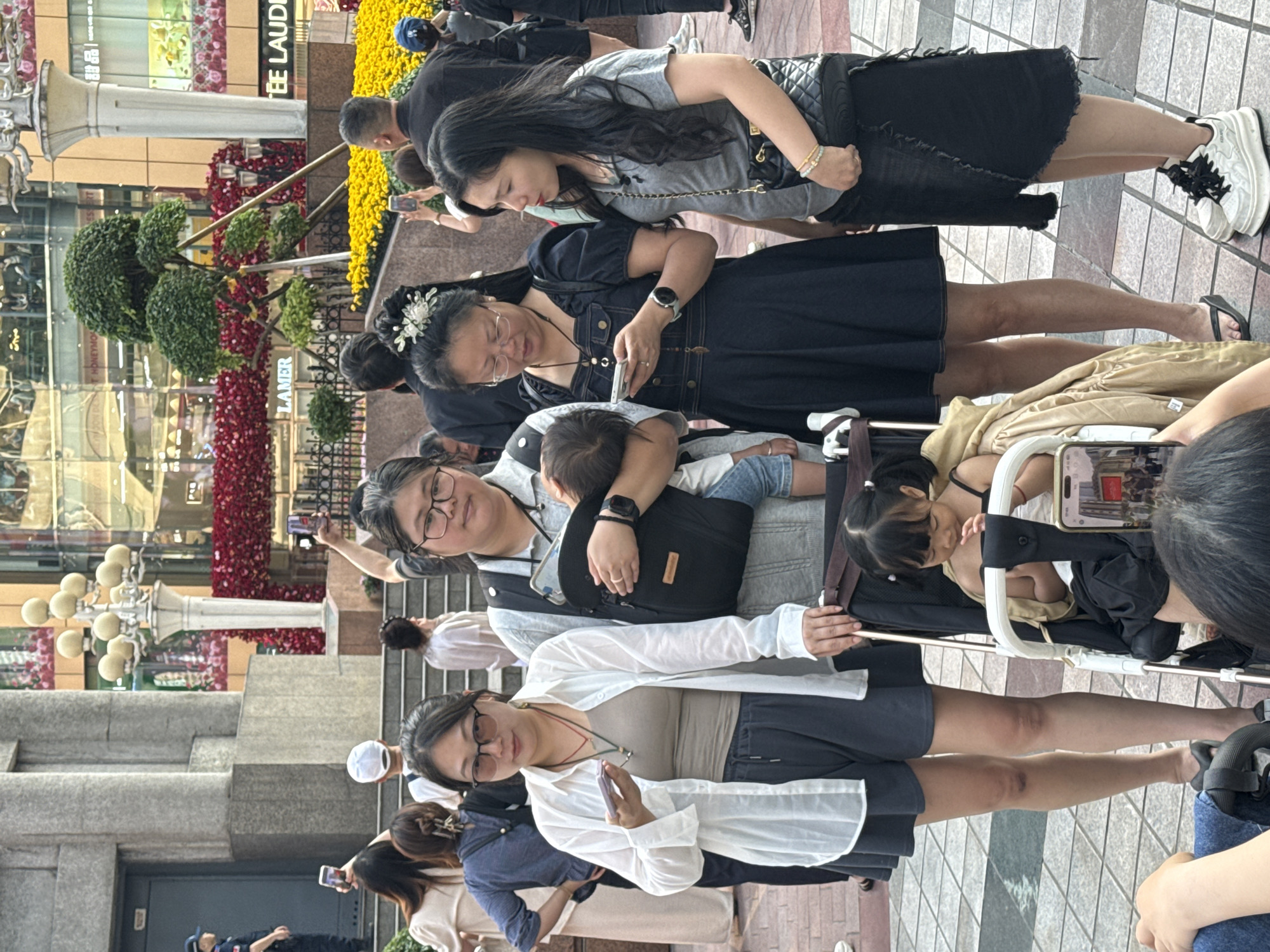 The image depicts a public space in the Yuzhong District of China, filled with people and decorative floral arrangements. Several individuals are seen holding and using mobile phones, suggesting they are taking photos or communicating. A woman is wearing a child carrier, holding a young child, and pushing a stroller. The floral decorations include red and yellow blossoms, arranged in a manner consistent with a public display or event. The overall environment suggests a busy urban area during daylight.