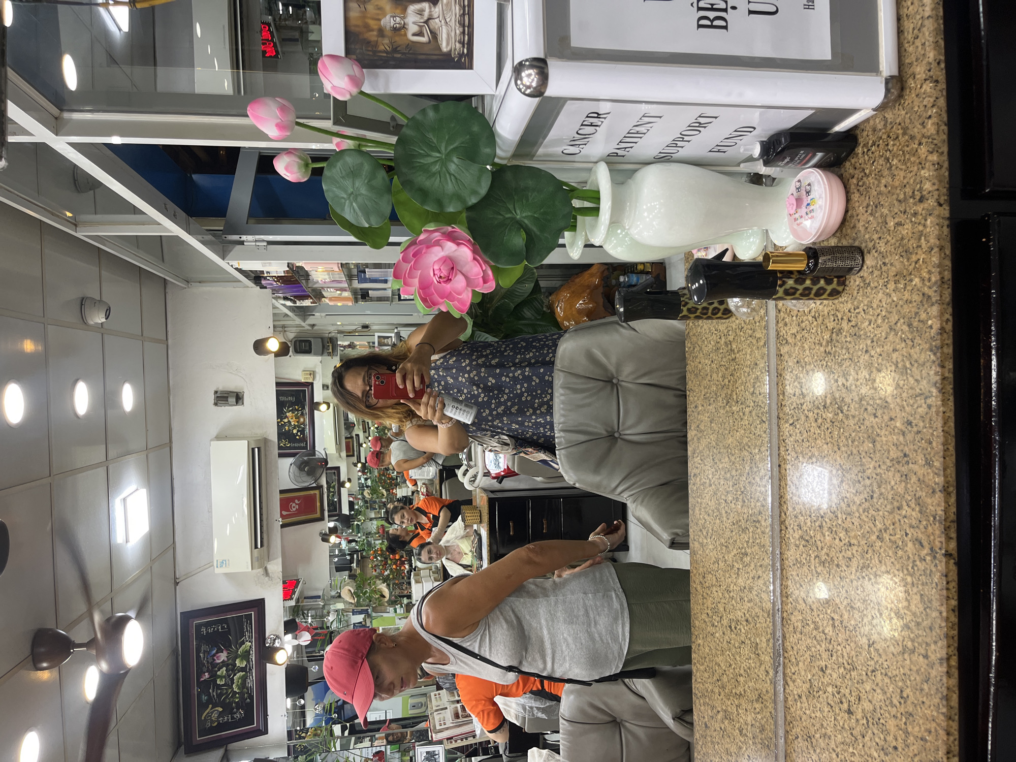 The image depicts the interior of a beauty salon in Nha Trang Ward, Vietnam. In the foreground, a mirror reflects a woman wearing a red baseball cap and a grey tank top, standing to the left. To the right, another woman, holding a red smartphone, is visible, appearing to take a selfie. She is wearing a patterned dress. A vase with artificial pink lotus flowers and green leaves is prominently placed on a counter in front of the mirror. Behind the counter, shelves filled with beauty products are visible. In the background, other individuals are seen working or being attended to. The salon appears well-lit with artificial lights and a ceiling fan overhead. The overall environment suggests a busy beauty salon setting.