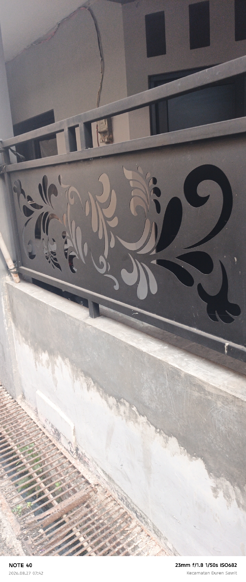 The image shows a close-up of a black metal fence with an ornate floral cutout design. The fence is positioned in front of a grey concrete wall. Below the fence, there is a concrete ledge that transitions to a rusted metal grate. The background consists of a building with a grey facade and dark rectangular windows. The lighting suggests it is daytime. Text at the bottom of the image indicates the date and time as "2026.08.27 07:42" and provides camera settings "23mm f/1.8 1/50s ISO682" along with the location "Kecamatan Duren Sawit", which is a subdistrict in East Jakarta, Indonesia.