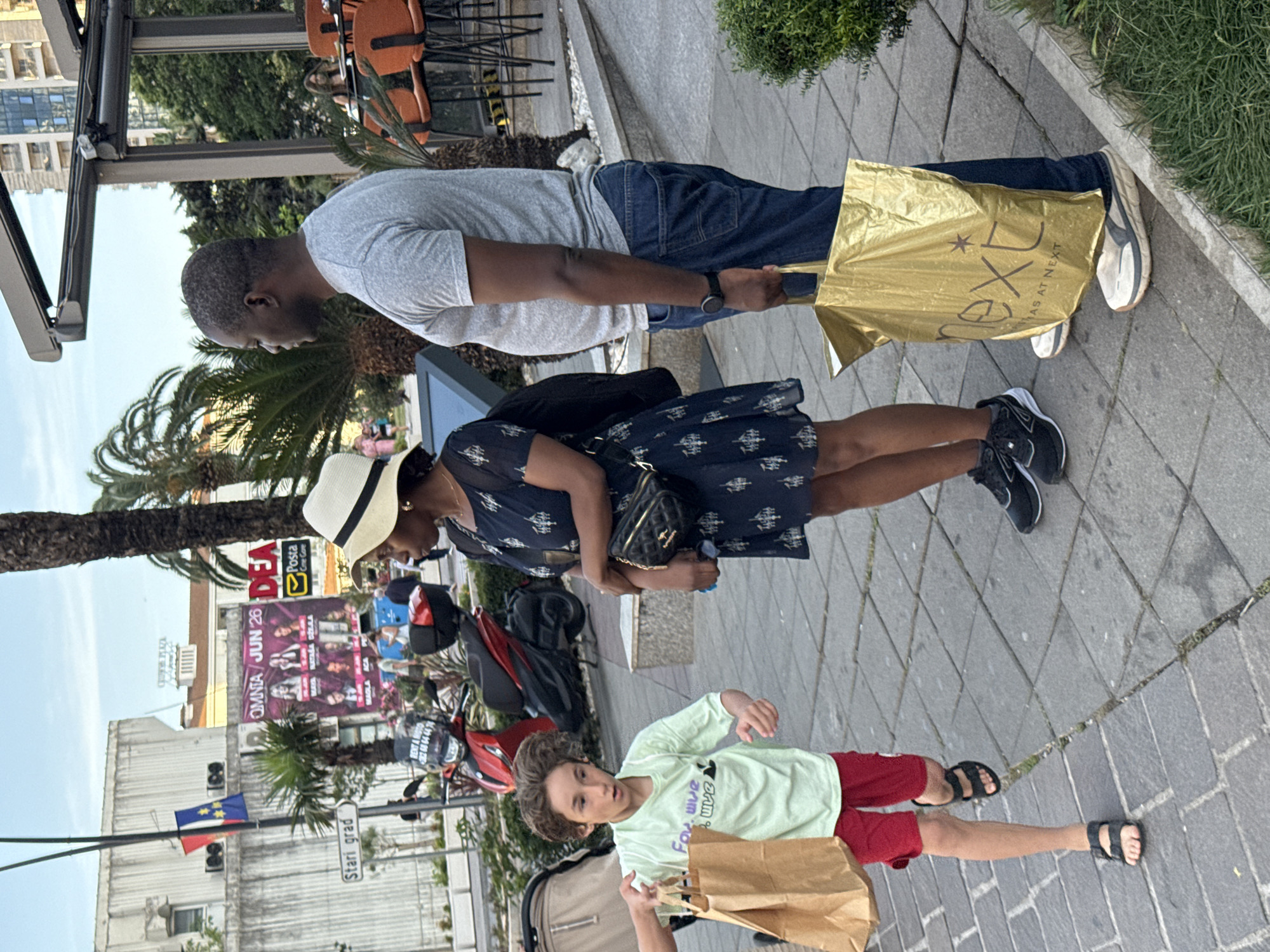A family of three is walking on a stone-tiled sidewalk in Budva, Montenegro. An African-American man in a grey t-shirt and jeans is carrying a gold shopping bag. A woman in a blue patterned dress and a straw hat is holding a black purse. A young boy in a green long-sleeved shirt and red shorts is carrying a brown shopping bag. In the background, there is a poster for "Omnia Jun 26" and signage for "Posta" and "Stari grad". A parked red motorcycle is visible, as well as a partial view of a building and palm trees. The shopping bag is from "next as at next".