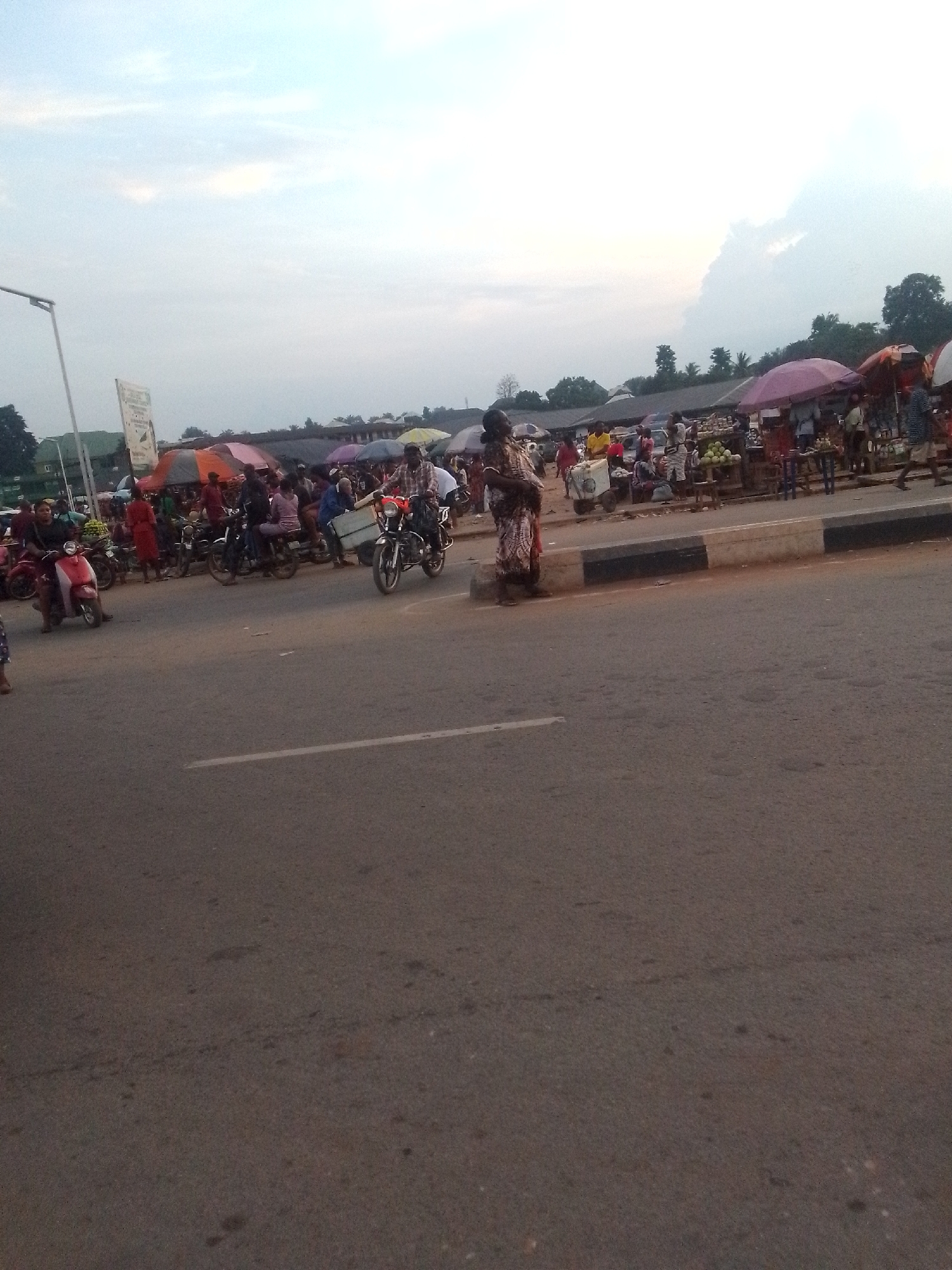 The image depicts a street scene, presumably in a market area in Unknown, Unknown. There are several motorcycles parked along the side of the road, some with people seated on them. A woman in a patterned dress stands on the edge of the road. Behind her and the motorcycles, numerous people are gathered, possibly engaged in commercial activities. Several stalls or stands with merchandise are visible, some covered by umbrellas. The sky is overcast, suggesting it might be early morning or late afternoon. The road itself occupies a significant portion of the image, appearing relatively empty.
