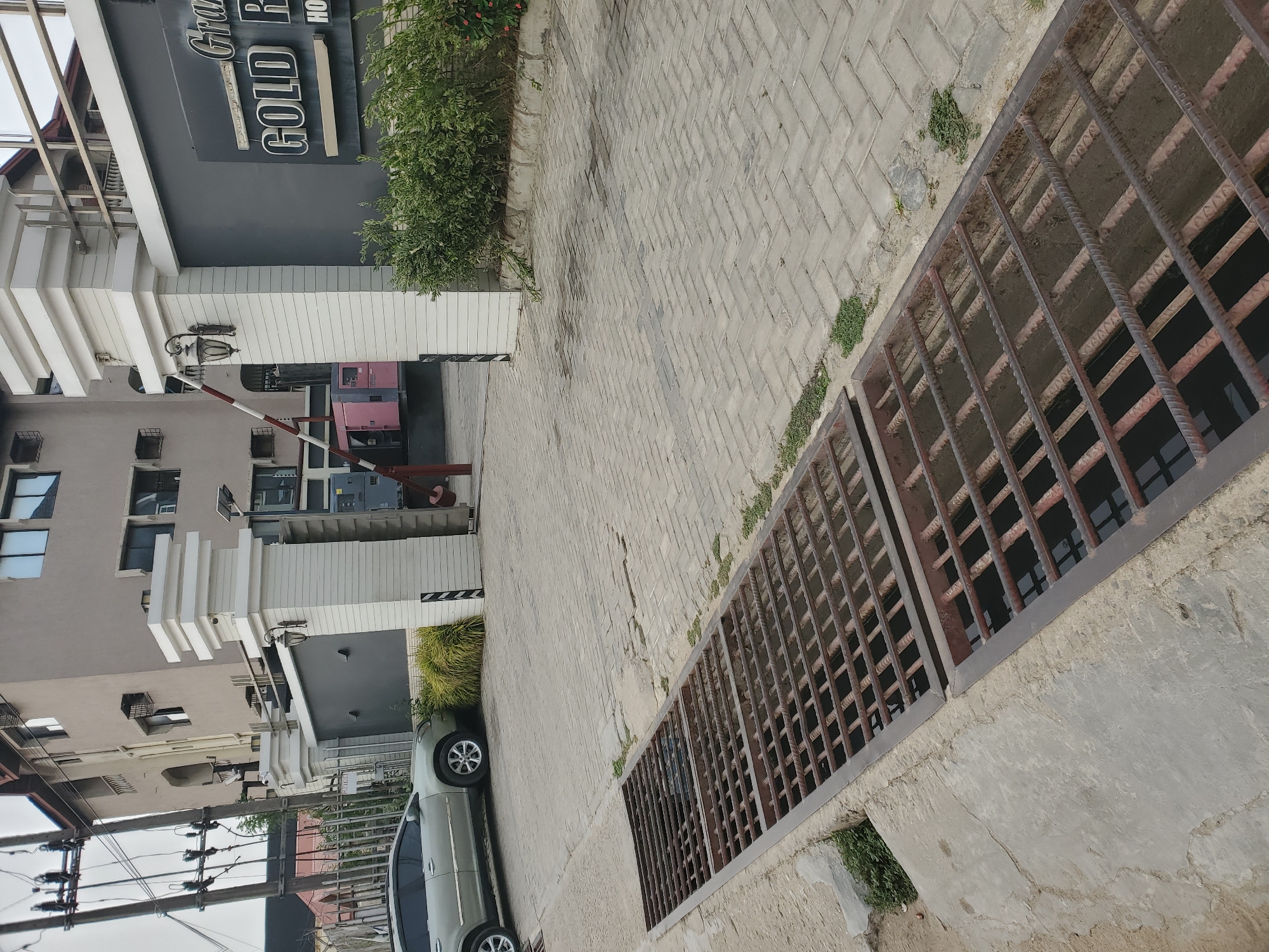 The image, taken in Lagos, Nigeria, captures an outdoor scene at the entrance of a commercial establishment, likely a hotel or resort. A multi-story building, predominantly grey and white with distinct architectural elements, occupies the left side of the frame. A dark sign on the building displays partial text: "Gra..." (likely "Grand"), "GOLD R", and "HO...", suggesting "Grand Gold Hotel" or "Resort".
At the building's entrance, a security checkpoint features a red and white boom gate, which is in a partially lowered position. Adjacent to the gate is a small security booth, and behind it, two prominent pink/magenta industrial generators are visible. A light-colored car, possibly a minivan or SUV, is parked to the right of the security booth. A person, partially obscured, can be seen near the boom gate.
The ground is paved with light-colored interlocking concrete bricks laid in a herringbone pattern, forming a driveway or walkway. In the foreground, a large rectangular metal grate, constructed from rusty rebar, covers an open drainage channel. Green bushes and small patches of grass grow along the edges of the paved areas and near the building. Utility poles with a network of electrical wires are visible on the far left of the frame. The scene is brightly lit, indicating daytime conditions.