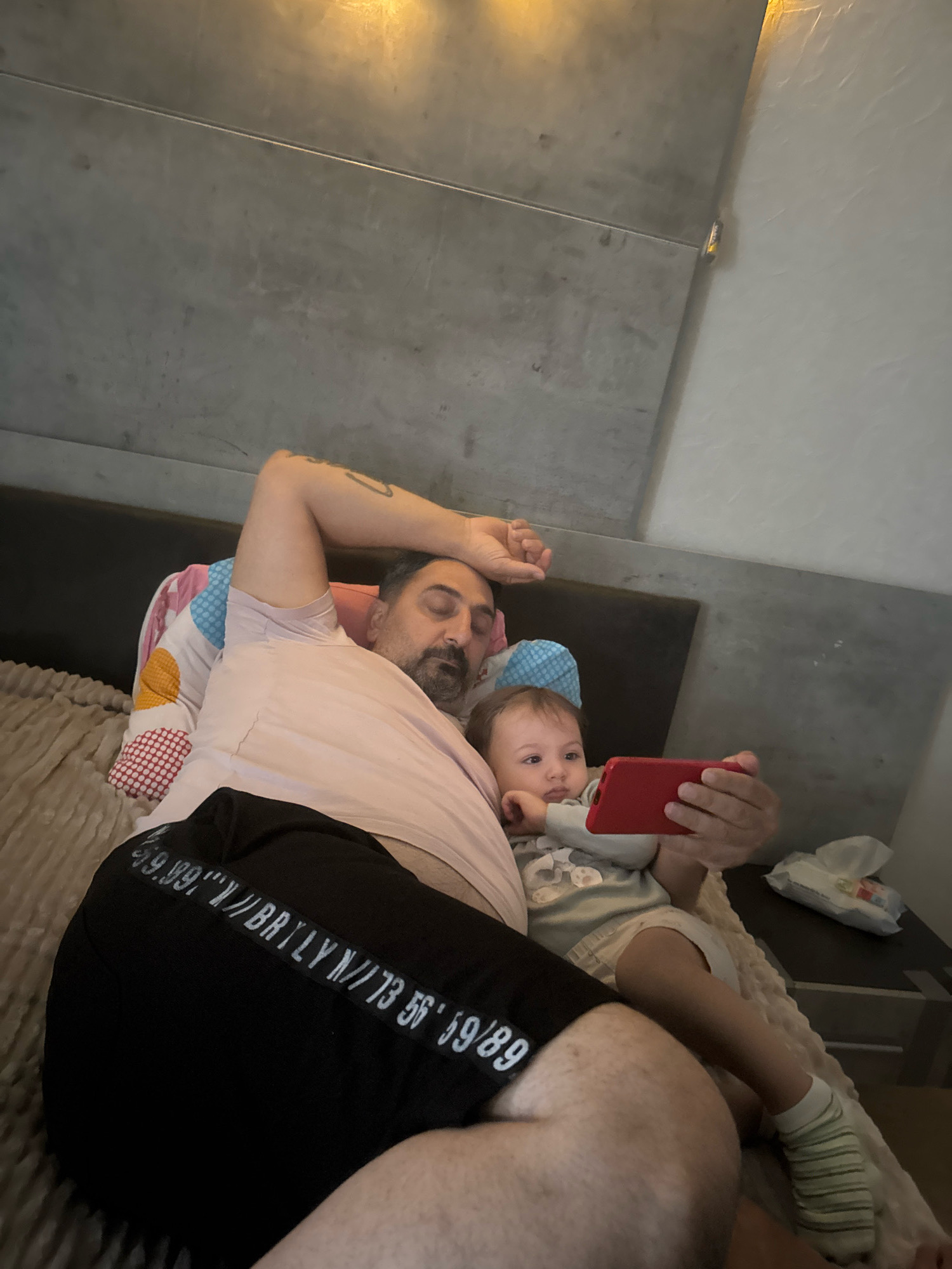 In Überherrn, Germany, a man and a baby are lying on a bed. The man, with dark hair and a beard, wears a light pink t-shirt and black shorts with white lettering. He is resting his head on a pillow and has his arm over his forehead, eyes closed. The baby, dressed in a light grey onesie, is sitting up and holding a red smartphone, looking at the screen. The bed has patterned pillows and is covered with a blanket. The background consists of a textured wall with metal accents and a bedside table with a pack of wipes. The scene appears to be an indoor domestic setting.