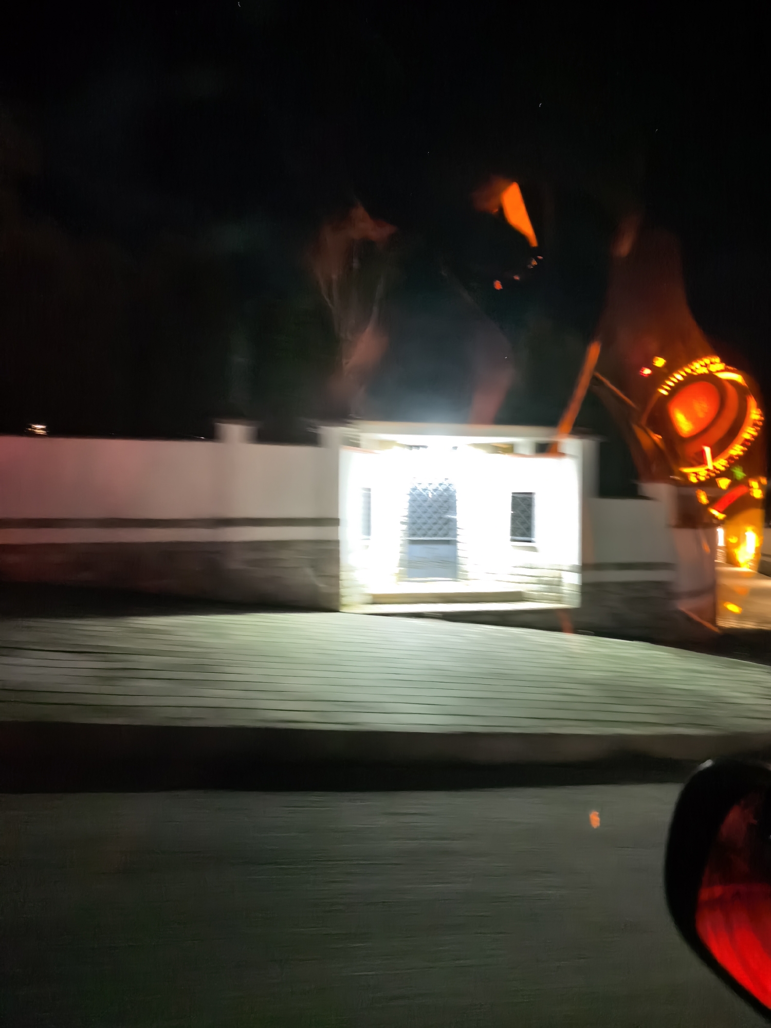 At night in an unknown location, a blurred image shows a white building with a fenced door and a window next to it, illuminated by a bright light. To the right of the building stands a large, orange illuminated structure with circular elements. In the foreground, a slightly reflective surface resembling a road or pavement is visible, with the edge of a car's red taillight on the right side of the image. The background is dark, with some tree-like forms visible behind the building. The image is blurred, suggesting motion.