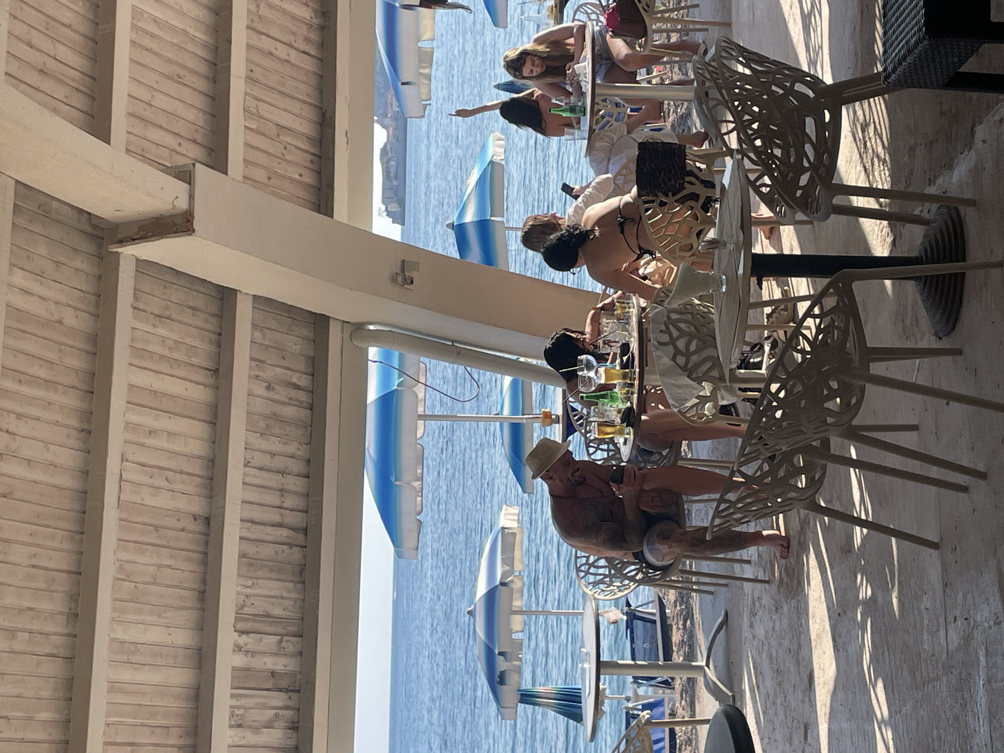 The image depicts a scene at a seaside restaurant in Budva, Montenegro. The view overlooks the Adriatic Sea, visible through a row of blue and white beach umbrellas. Several groups of people are seated at tables, some dining and others relaxing. A man wearing a hat is prominent in the foreground, engaged with his phone. The restaurant has a roof with a wooden structure. Tables and chairs, some of which are of a modern, ornate design, are arranged on a stone-paved area. The lighting suggests it is daytime, with strong sunlight casting shadows on the ground.