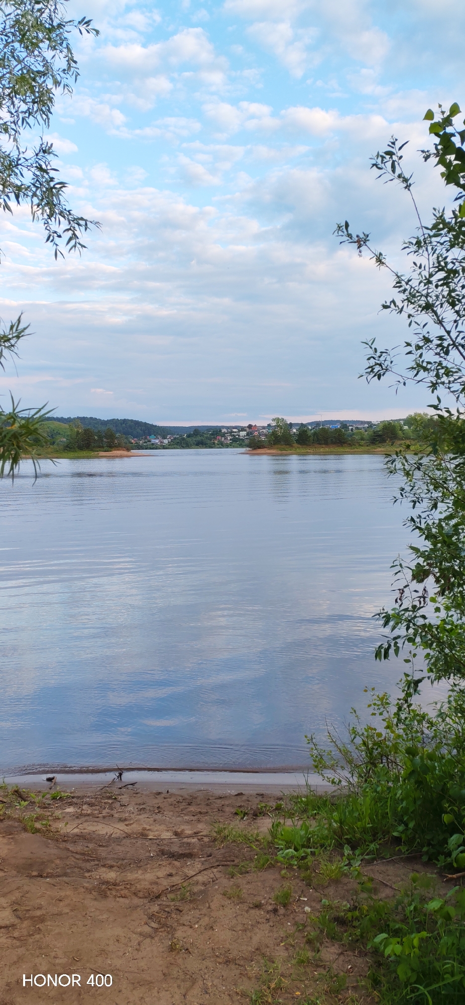 The image depicts a serene natural landscape with a wide, calm body of water in the foreground, reflecting the sky and clouds. On the far bank of the water, a small town or village is visible, nestled among rolling hills covered in trees. The architecture of the buildings suggests a rural or semi-rural setting. The sky is partly cloudy with a soft, diffused light, indicating it might be late afternoon or early morning. The foreground shows a sandy bank with some vegetation and branches, suggesting a natural shoreline. The overall atmosphere is peaceful and tranquil. The location is identified as Noviy, Russia.