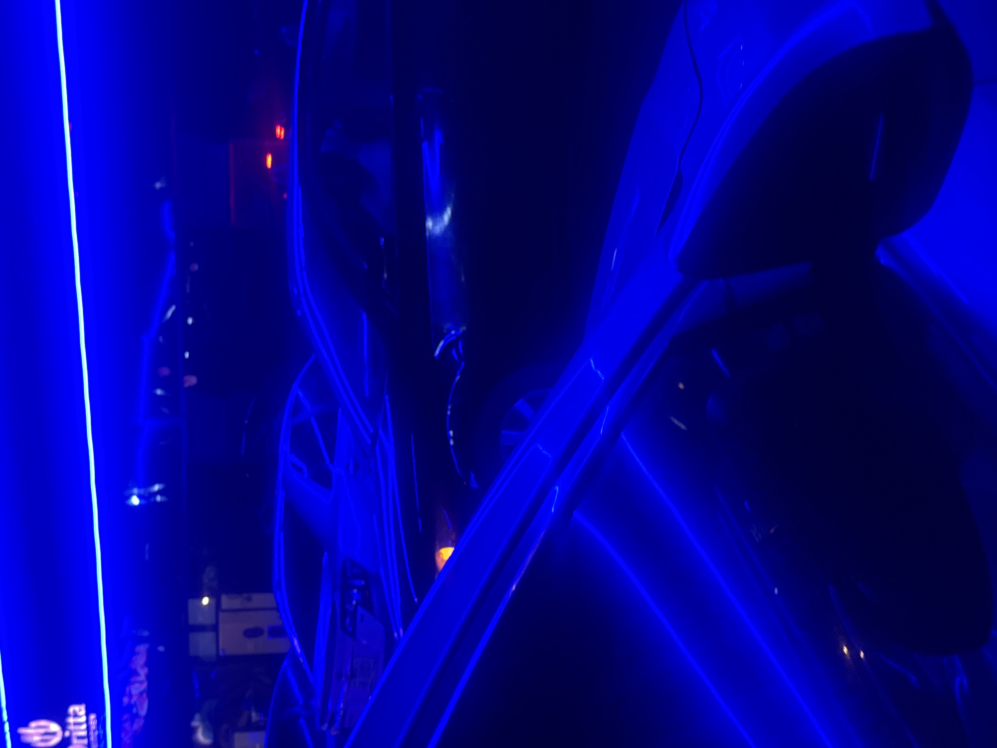 The image depicts a nighttime scene with prominent blue neon lighting. In the background, a sign with the word "Dritta" is visible, illuminated by blue lights, suggesting it might be a business or establishment. Several cars are parked, their shapes partially discernible due to the low light and the reflections from the blue neon. One car in the foreground has its doors open, with a visible interior, also bathed in blue light. The overall impression is that of an urban environment, potentially a parking lot or street in a nightlife district. The location of the city of Abuja, Nigeria, is not discernible from the visual information in the image itself.