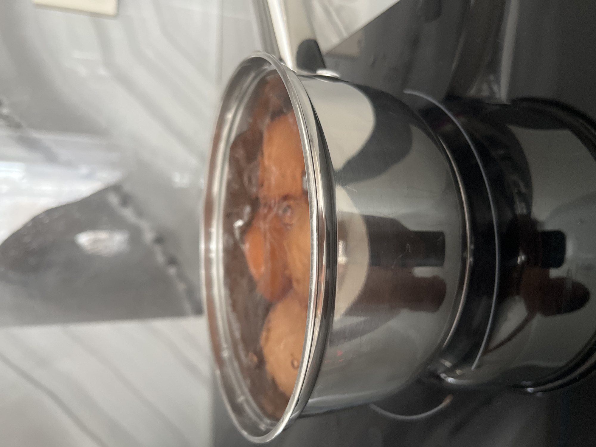 A close-up, eye-level shot shows a stainless steel pot filled with boiling water and several brown eggs. The pot is on a black electric stove burner. Steam rises from the pot, obscuring the background, which appears to be a light-colored tiled wall with a subtle pattern. The lighting is bright, highlighting the metallic sheen of the pot and the bubbles in the water. No people are visible in the frame. The scene is likely from a kitchen environment. The location is not specified in the image or its crops.