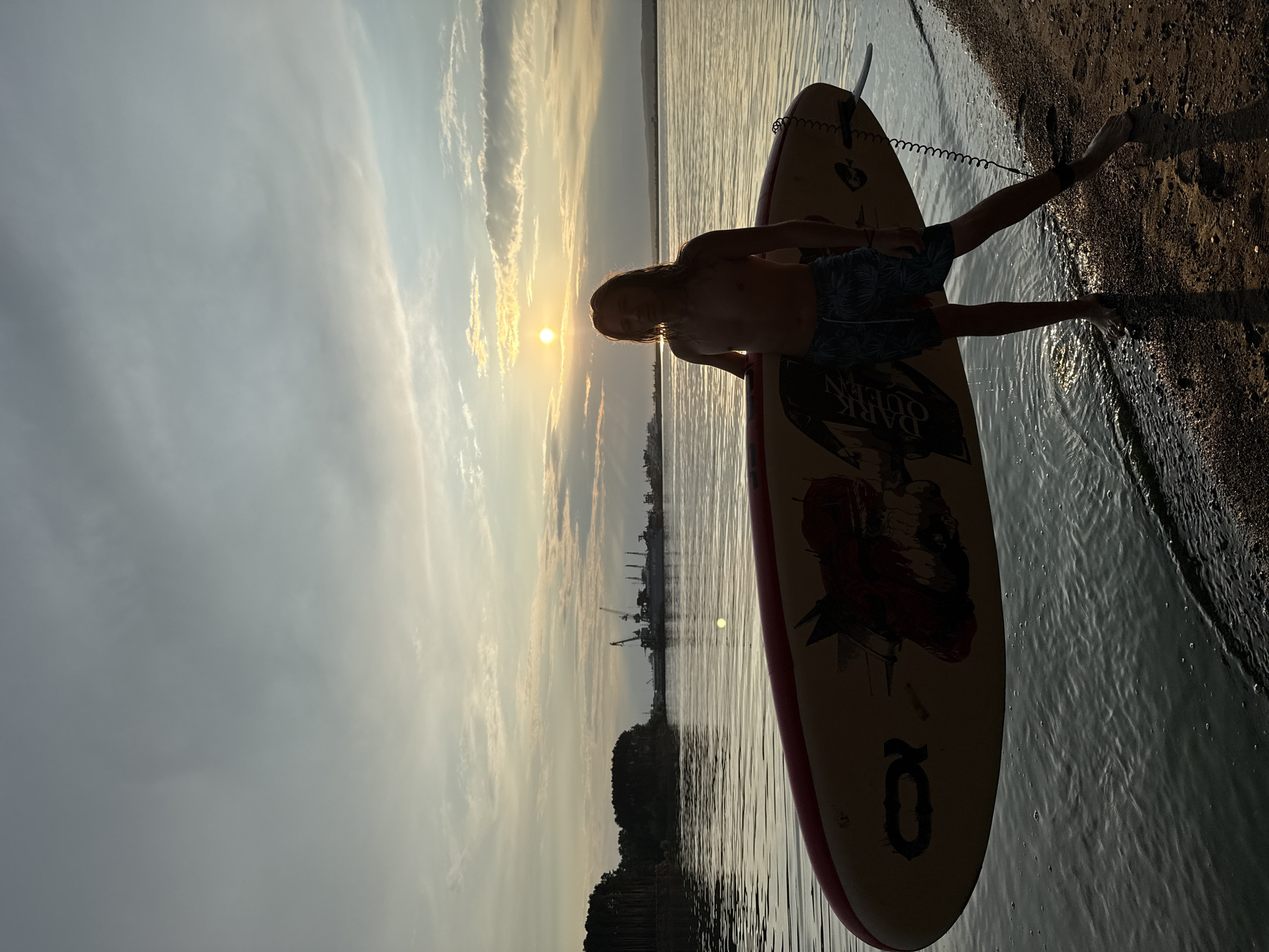A young man with long blonde hair, wearing patterned swim trunks, stands on a sandy beach at the edge of a calm body of water. He is shirtless and holds a paddleboard horizontally in front of him. The paddleboard is yellow with red trim and features graphics. The sun is setting or rising, casting a warm glow and silhouetting the man and the distant treeline and industrial structures across the water. The sky is filled with scattered clouds, and the water reflects the light of the sky. The scene appears to be a serene natural or semi-industrial waterfront at either dawn or dusk.