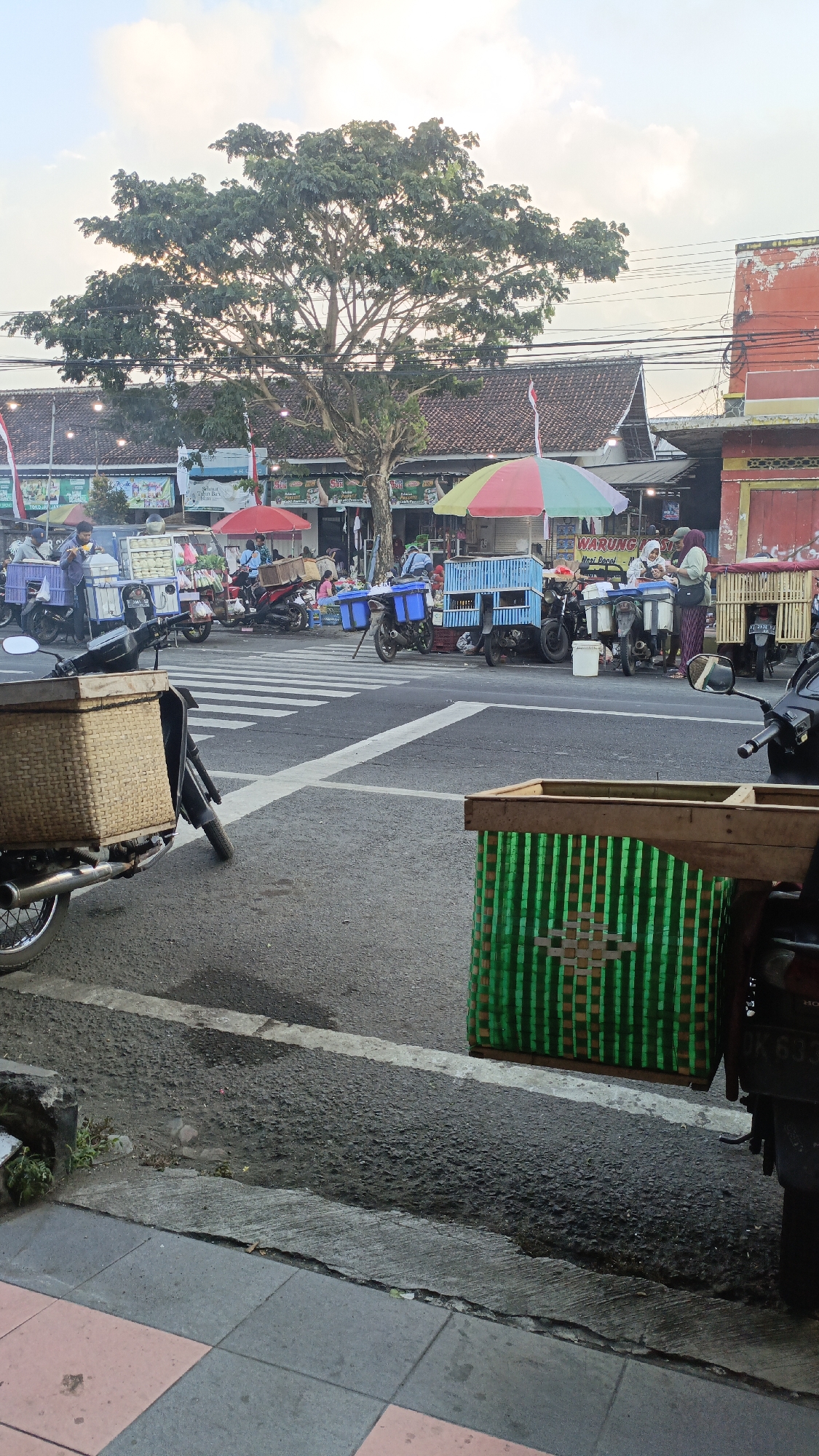 The image captures a street scene in Srono, Indonesia, likely a bustling market area near a road intersection. In the foreground, two motorcycles are parked. The one on the left has a large, woven wicker basket attached to its rear. The motorcycle on the right has a green woven basket with a wooden frame mounted on its rear. The road is paved and marked with white pedestrian crossing lines.
In the background, several food vendors are set up along the side of the road, many operating from motorcycles or small carts. They are sheltered by colorful umbrellas, including a prominent red, yellow, and green one. People are visible interacting with the vendors, some purchasing food, and others standing around. The vendors appear to be selling various items from their carts, some of which are blue and rectangular, possibly for carrying goods. In the far background, buildings with tiled roofs line the street, and a large tree with dense foliage stands prominently, casting a shadow over the scene. The sky is partly cloudy, suggesting it might be late afternoon or early morning. The overall atmosphere is one of daily commerce and local activity.