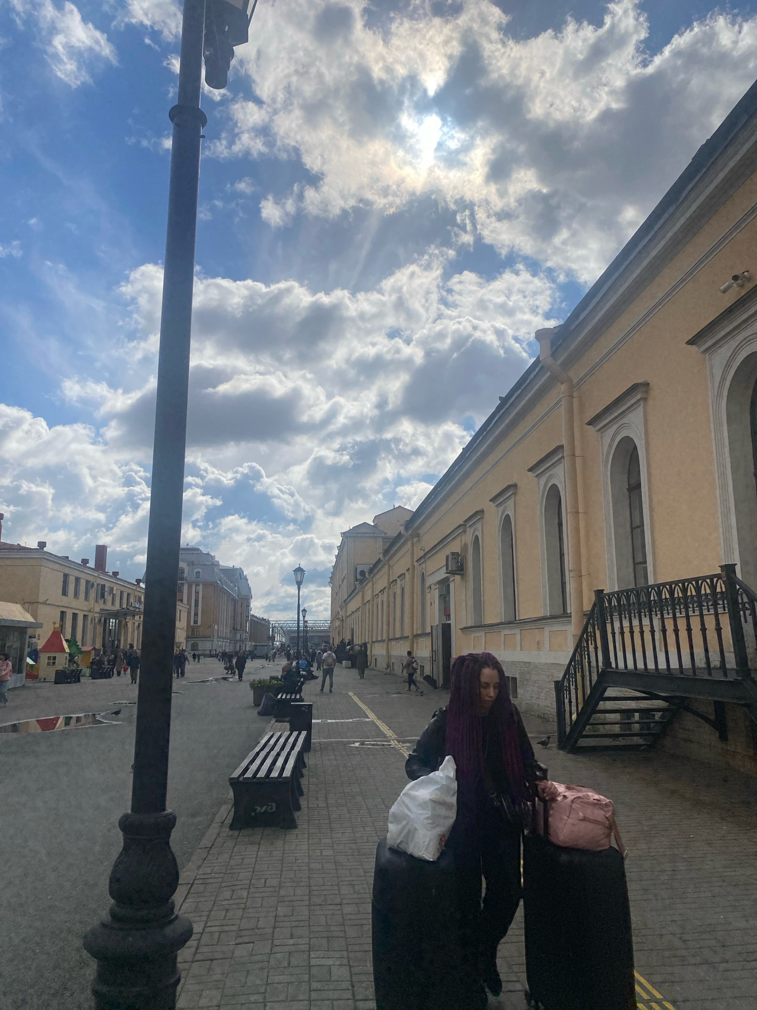 In Saint Petersburg, Russia, a woman with dark, purple dreadlocks walks along a cobblestone sidewalk next to a yellow building with arched windows. She is pulling two black suitcases and carrying a white plastic bag in her left hand and a pink duffel bag on top of one of the suitcases. The sun shines through a cloudy sky, casting shadows on the ground. A black lamppost stands to the left of the woman, and benches are visible along the sidewalk. In the background, people are walking, and more buildings line the street, with a train track structure in the distance. The scene appears to be a train station or a similar public transportation hub.