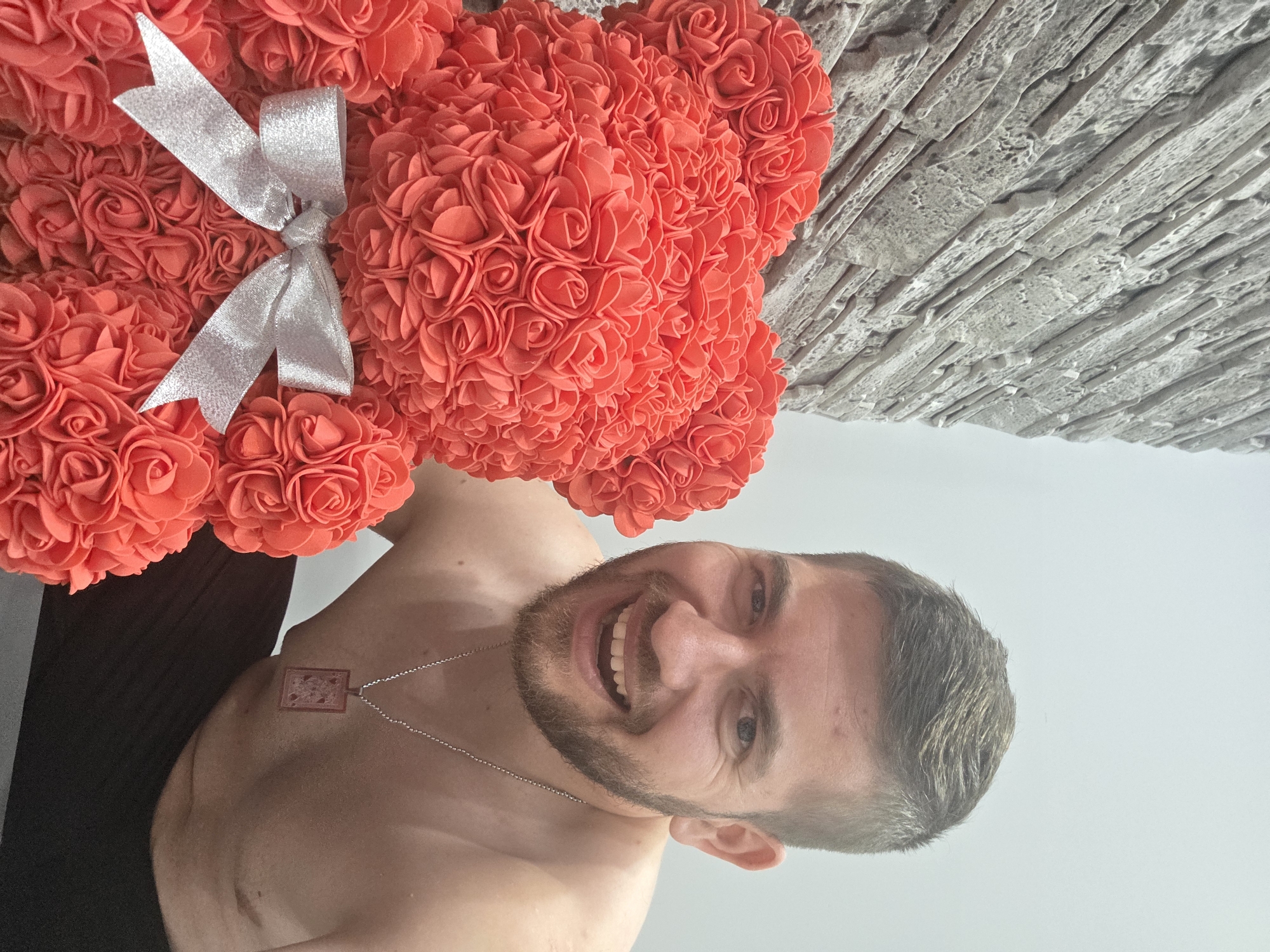 A man with a short haircut and a beard is smiling at the camera while holding a red teddy bear made of roses. The teddy bear has a silver bow around its neck. The man is wearing a silver chain necklace. The background behind the man is a textured grey wall. The picture was taken indoors, in a location unknown, outside of Yagodovo, Bulgaria.