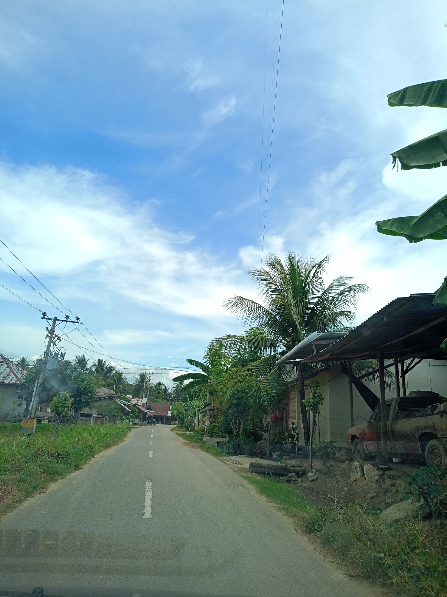The image depicts a road in Seuneuboklhong, Indonesia. The road stretches into the distance, flanked by greenery and buildings. Power lines run above the road. The sky is blue with scattered clouds. On the right, a dilapidated building with a rusted truck parked beneath its shelter is visible. Vegetation is lush.
