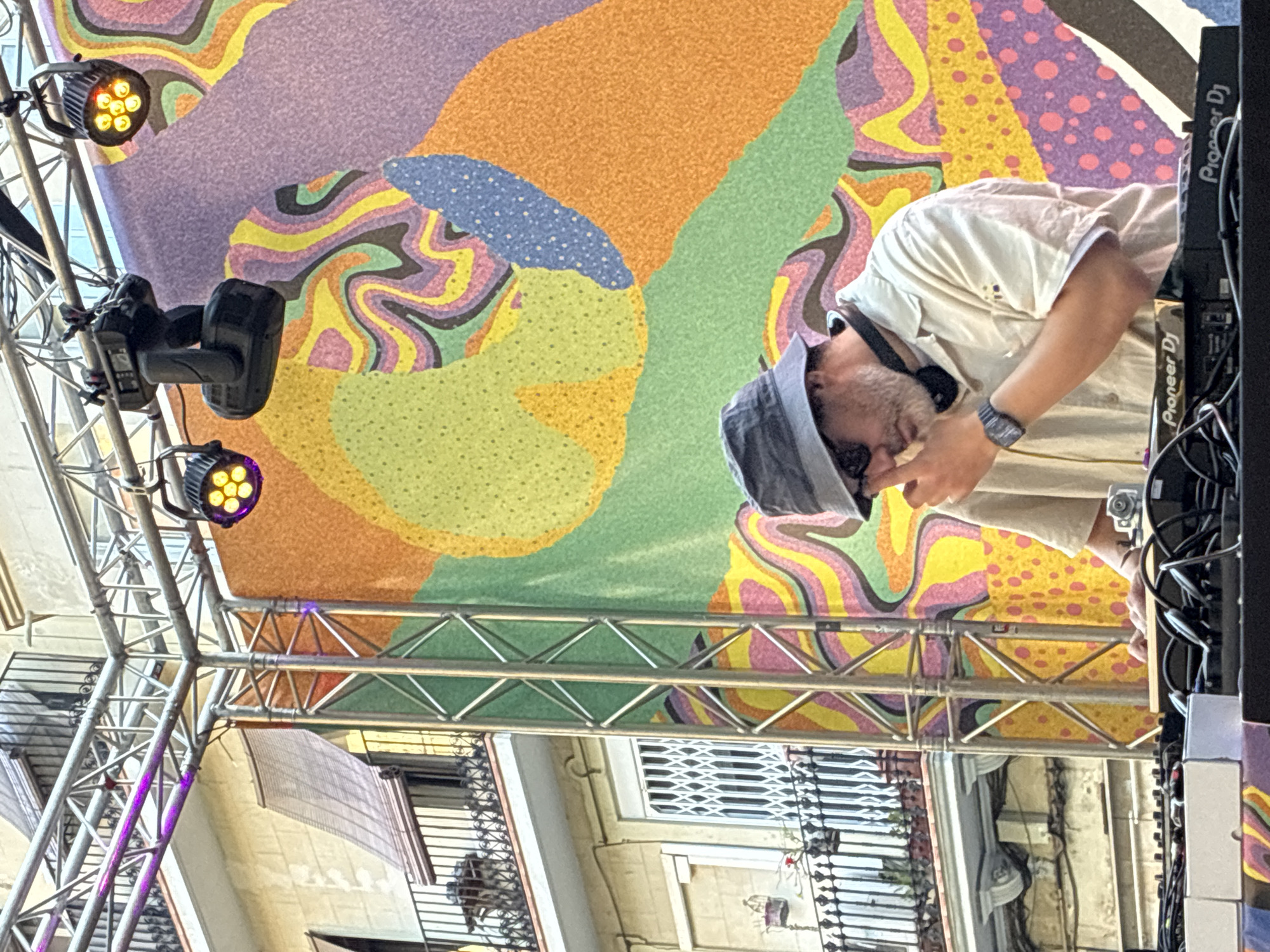 The image shows a DJ performing at an outdoor event, possibly in Barcelona, Spain, given the architecture. The backdrop features a colorful, abstract mural in purples, oranges, greens, and yellows. A lighting rig with multiple fixtures is visible in the foreground. The DJ, wearing a hat and sunglasses, is operating Pioneer DJ equipment. The event appears to be taking place in an urban area with buildings visible in the background.