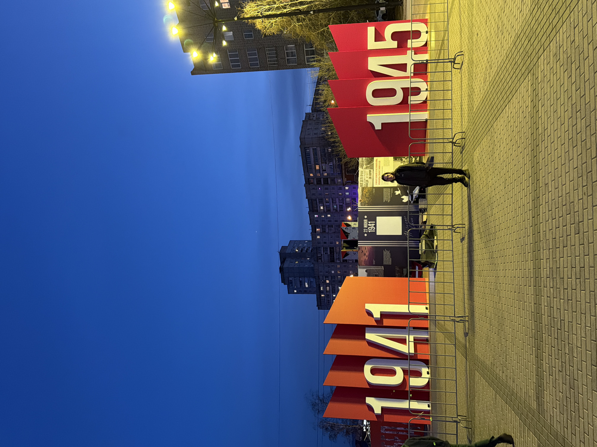 The image depicts an outdoor exhibit, likely commemorating World War II, in the city of Krasnoyarsk, Russia. Large red placards display the numbers "1941" and "1945" in white, indicating the start and end years of the war.
A person in dark clothing stands near the "1945" placard. The exhibit is bordered by metal barriers. Informational panels with text in Russian and historical images are visible.
The background includes residential buildings and streetlights, suggesting an urban setting. The sky is a deep blue, indicating either dusk or dawn. The ground is paved with rectangular stones.